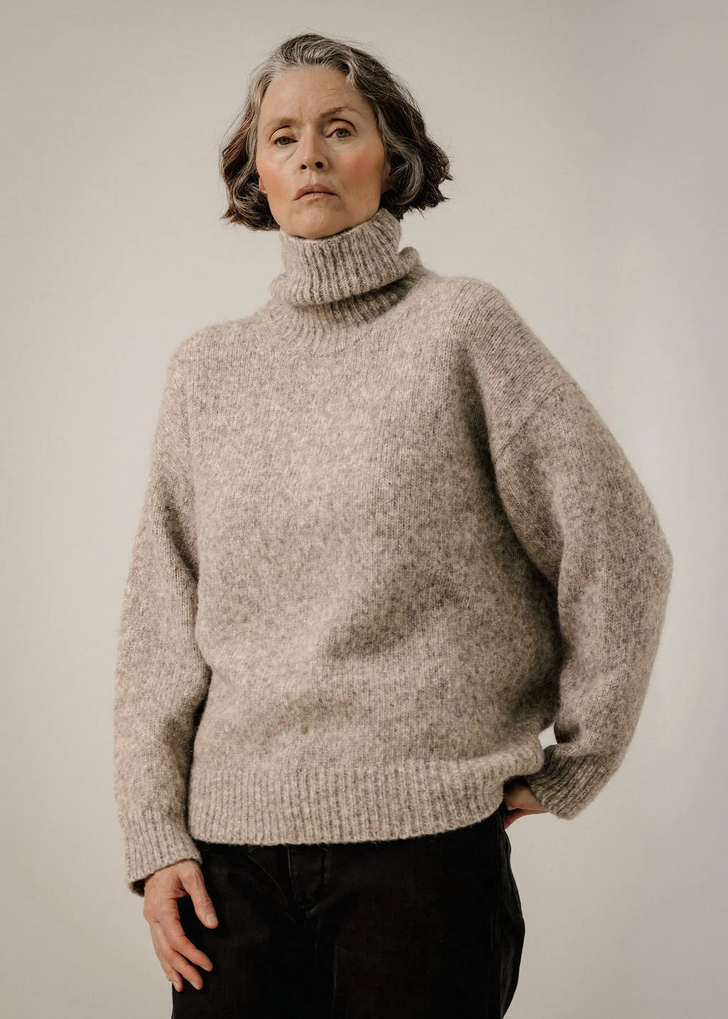 A woman wearing a chunky, oversized turtleneck sweater in a heathered light taupe-beige color, paired with black pants, photographed against a neutral light gray studio background.