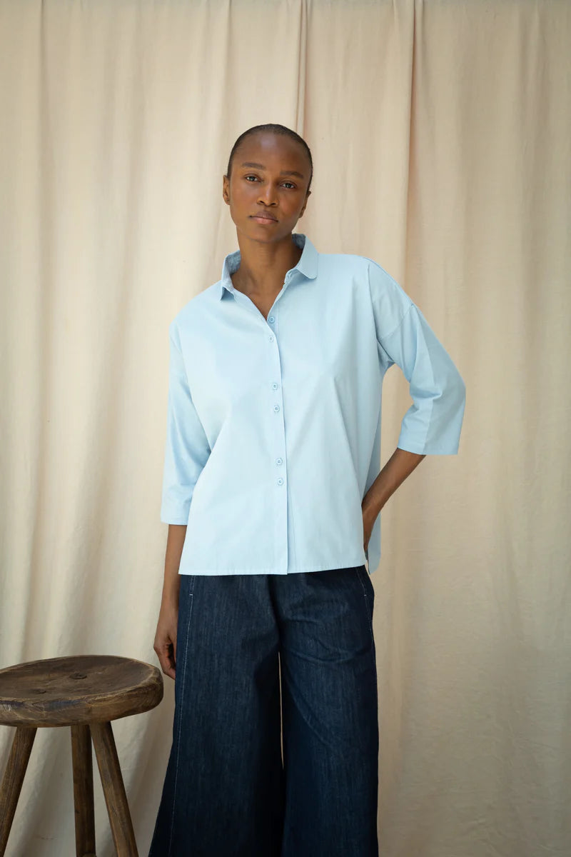 Person wearing a light blue shirt and dark pants standing against a beige curtain.