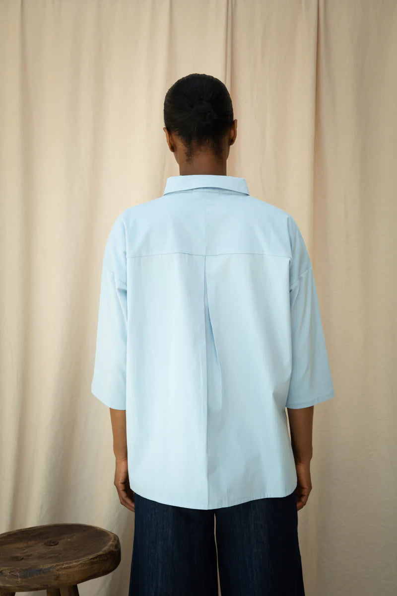 Person wearing a light blue shirt standing against a beige curtain