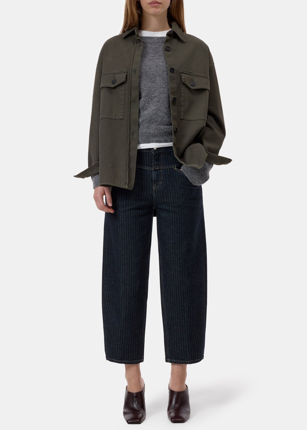 A model is shown from the neck down wearing an olive green utility jacket over a gray knit sweater, paired with dark navy pinstriped cropped jeans and dark brown leather ankle boots, against a light gray studio background.