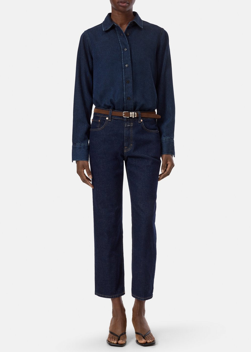 A model wearing a dark indigo denim long-sleeve button-up shirt tucked into matching dark blue jeans, styled with a brown leather belt and brown sandals, against a plain light gray background.