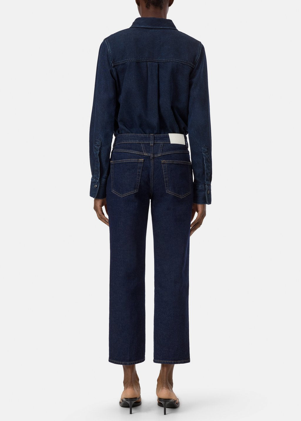 A model viewed from the back wearing a dark indigo denim set consisting of a long-sleeve button-up shirt and matching cropped wide-leg jeans, paired with black pointed-toe heels against a light gray studio background.