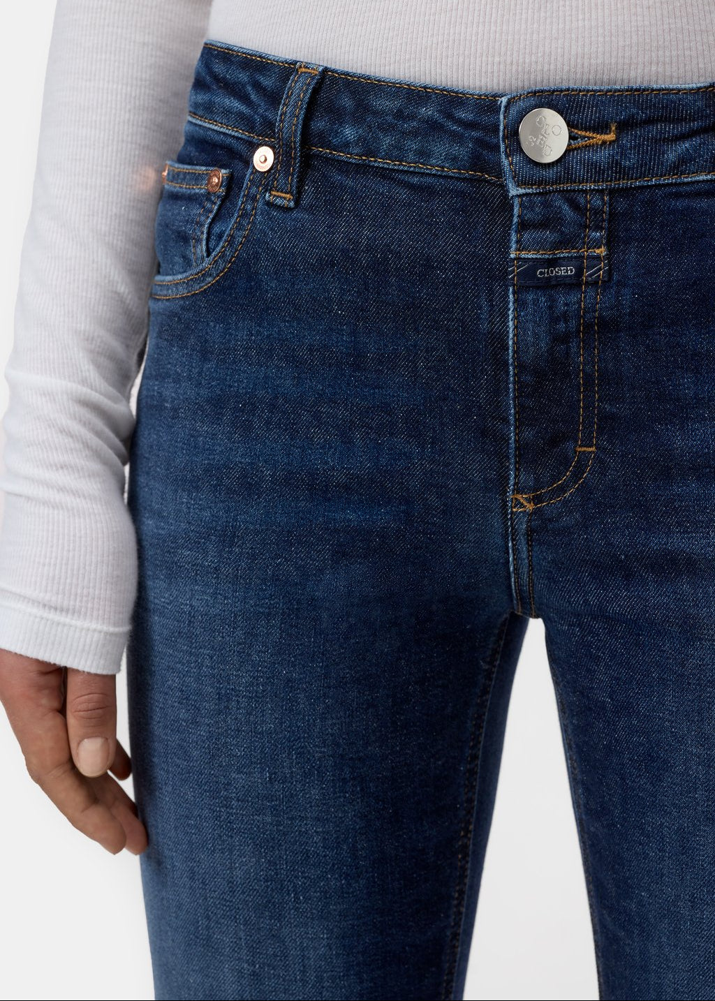Close-up of a person wearing dark blue denim jeans with a white ribbed long-sleeve top, showing the front waistband, button, belt loops, and pocket details.