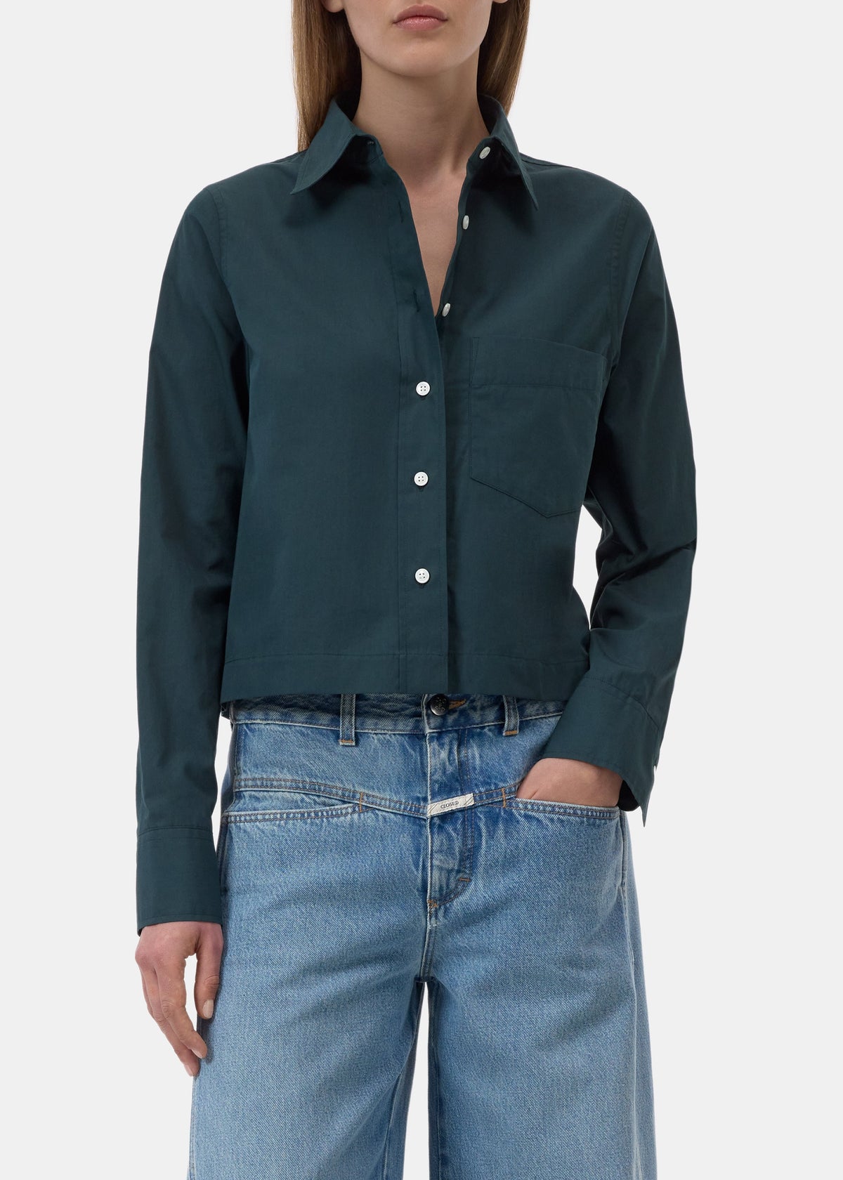 A woman wearing a dark teal-green long-sleeve button-up shirt with a chest pocket, paired with light-wash blue jeans, shown from the neck down against a light gray background.