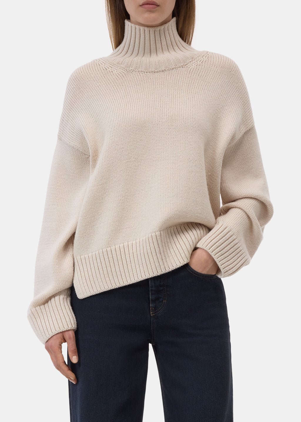 A woman wearing a cream-colored ribbed knit turtleneck sweater and dark navy blue jeans, shown from the neck down against a plain light gray background.