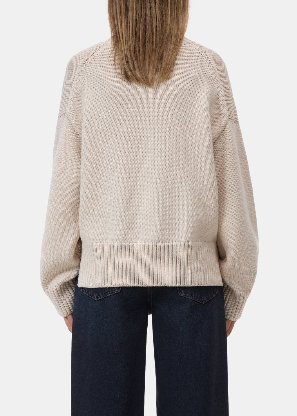 A person shown from the back wearing a light beige knit sweater with ribbed cuffs and hem, paired with dark navy pants, against a plain light gray studio background.