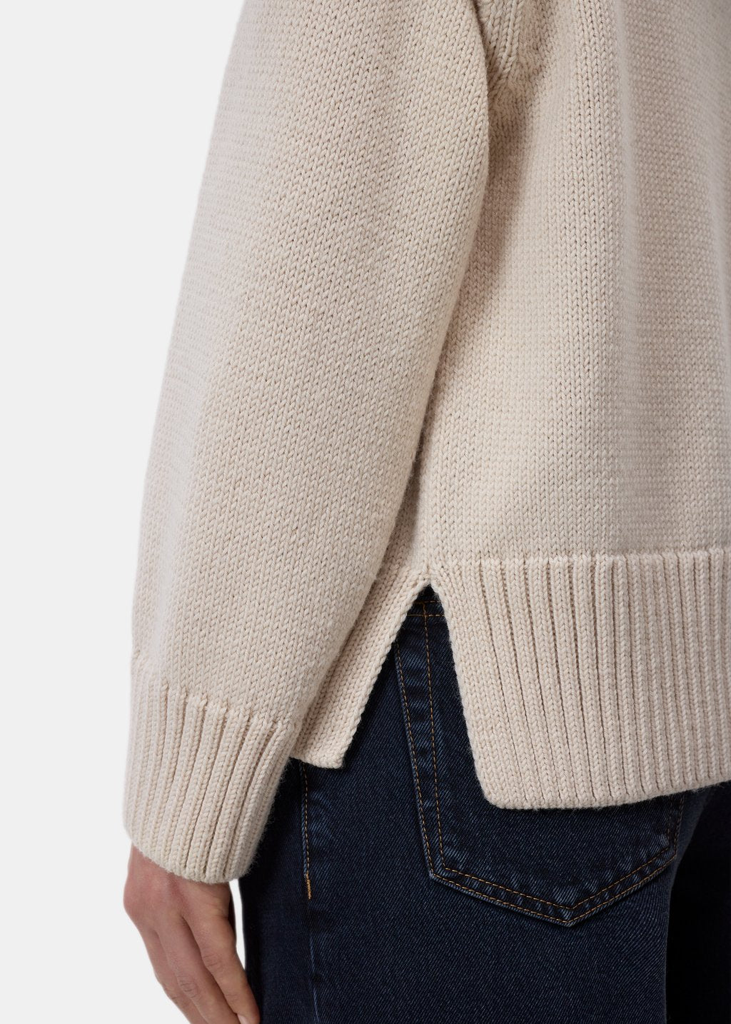 Close-up of a person wearing a cream-colored knit sweater with dark blue jeans, showing the ribbed cuffs and hem against a light gray background.