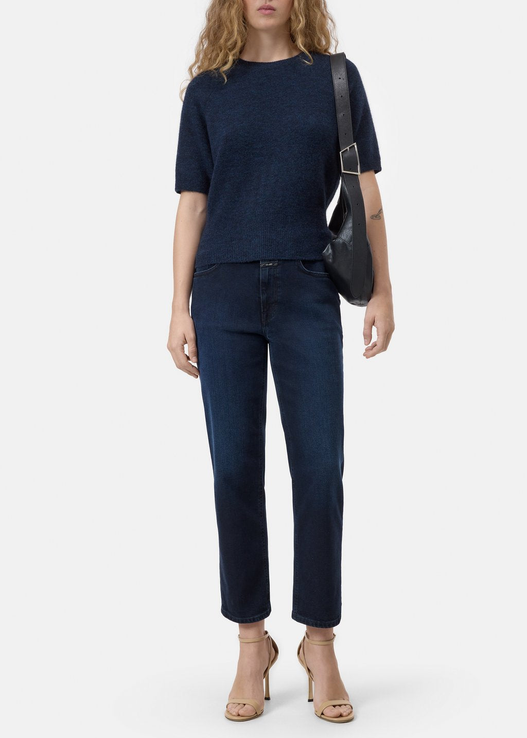 A model wearing a dark navy blue short-sleeve knit top and matching dark blue cropped jeans, styled with a black shoulder bag and beige high-heeled sandals against a light gray studio background.