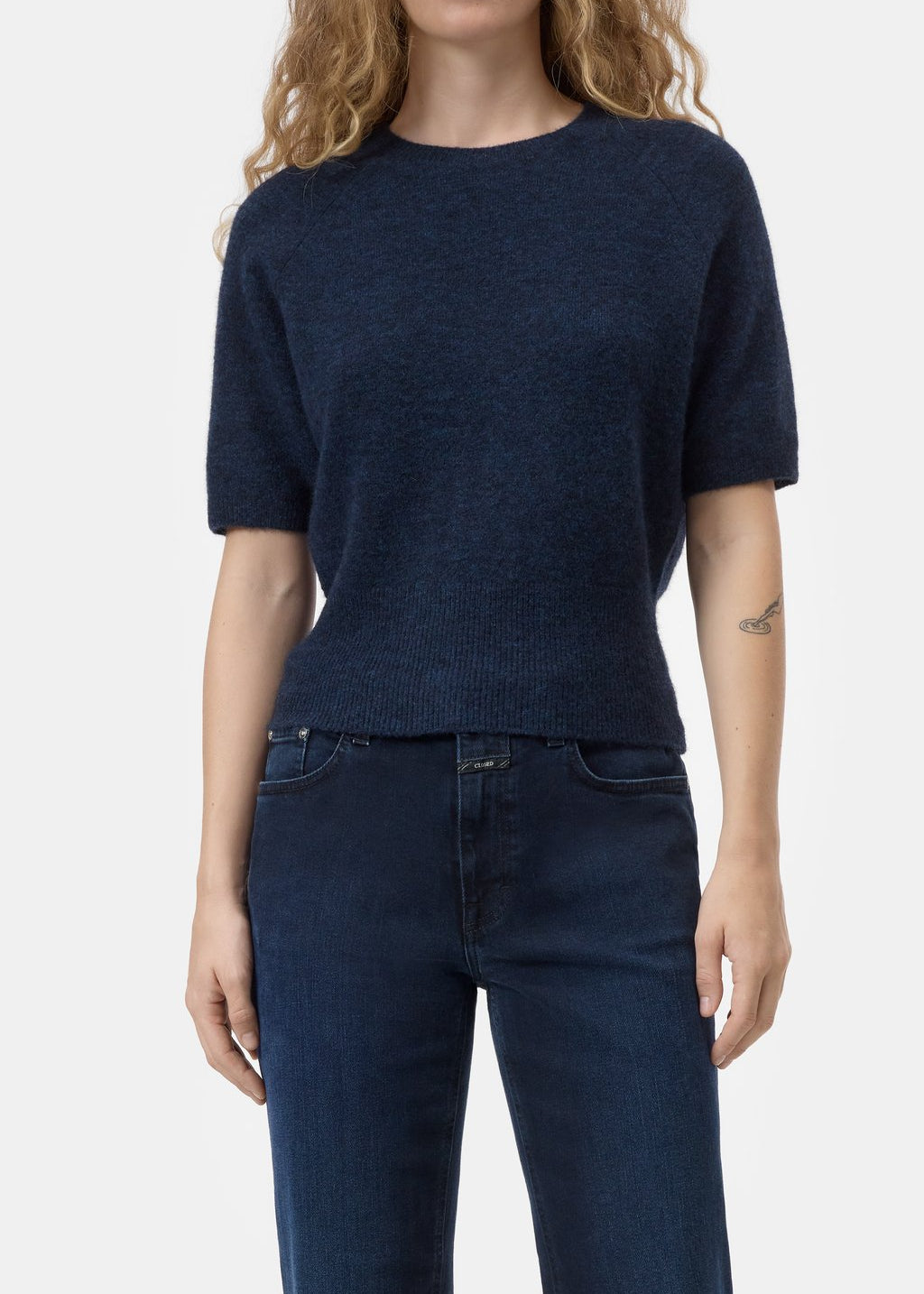 A woman wearing a dark navy blue short-sleeve knit sweater and dark blue jeans, shown from the neck down against a light gray studio background.