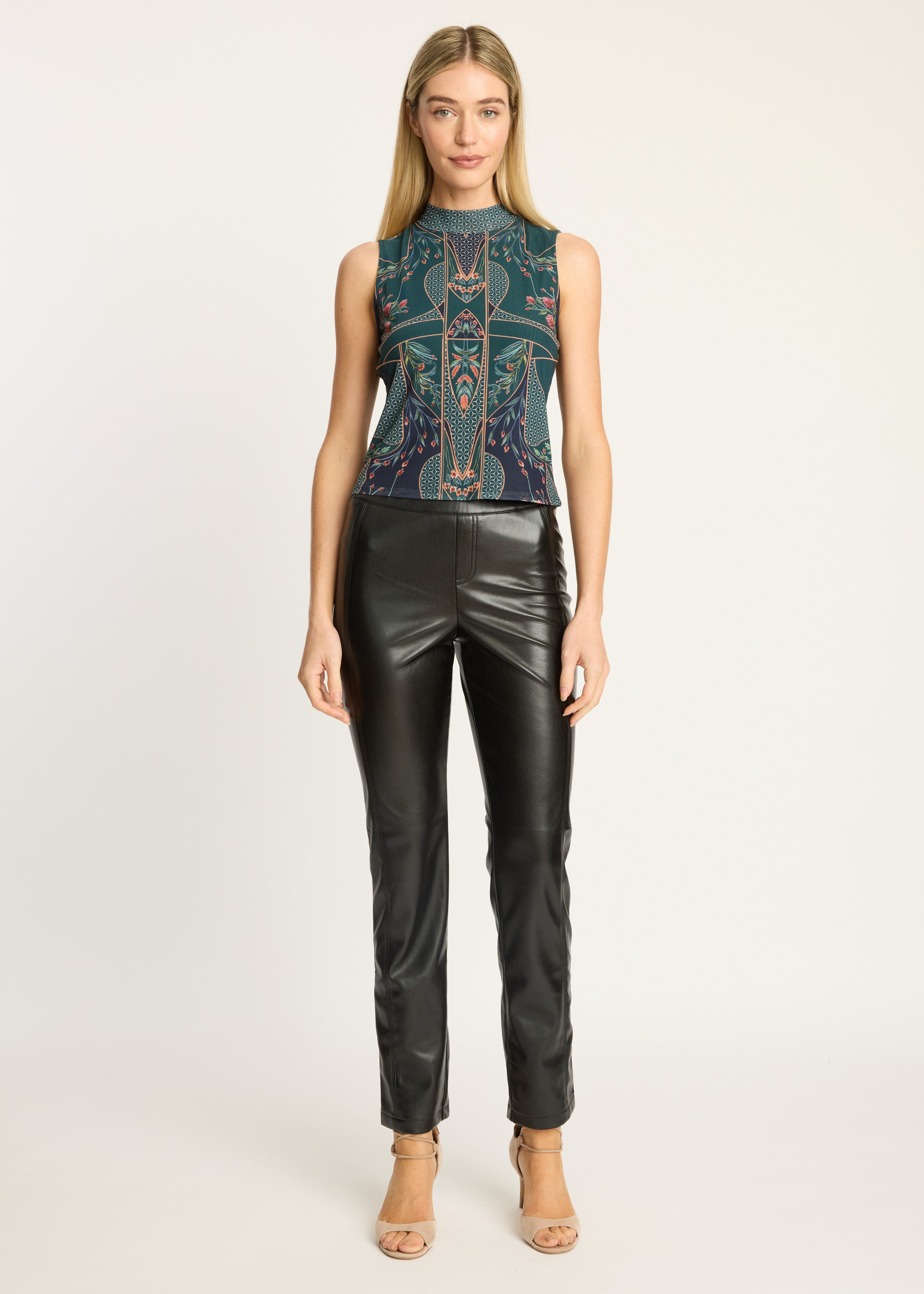 A woman modeling a sleeveless mock-neck top with a dark teal and black geometric pattern, paired with black faux-leather pants and beige high-heeled sandals, against a plain light gray studio background.