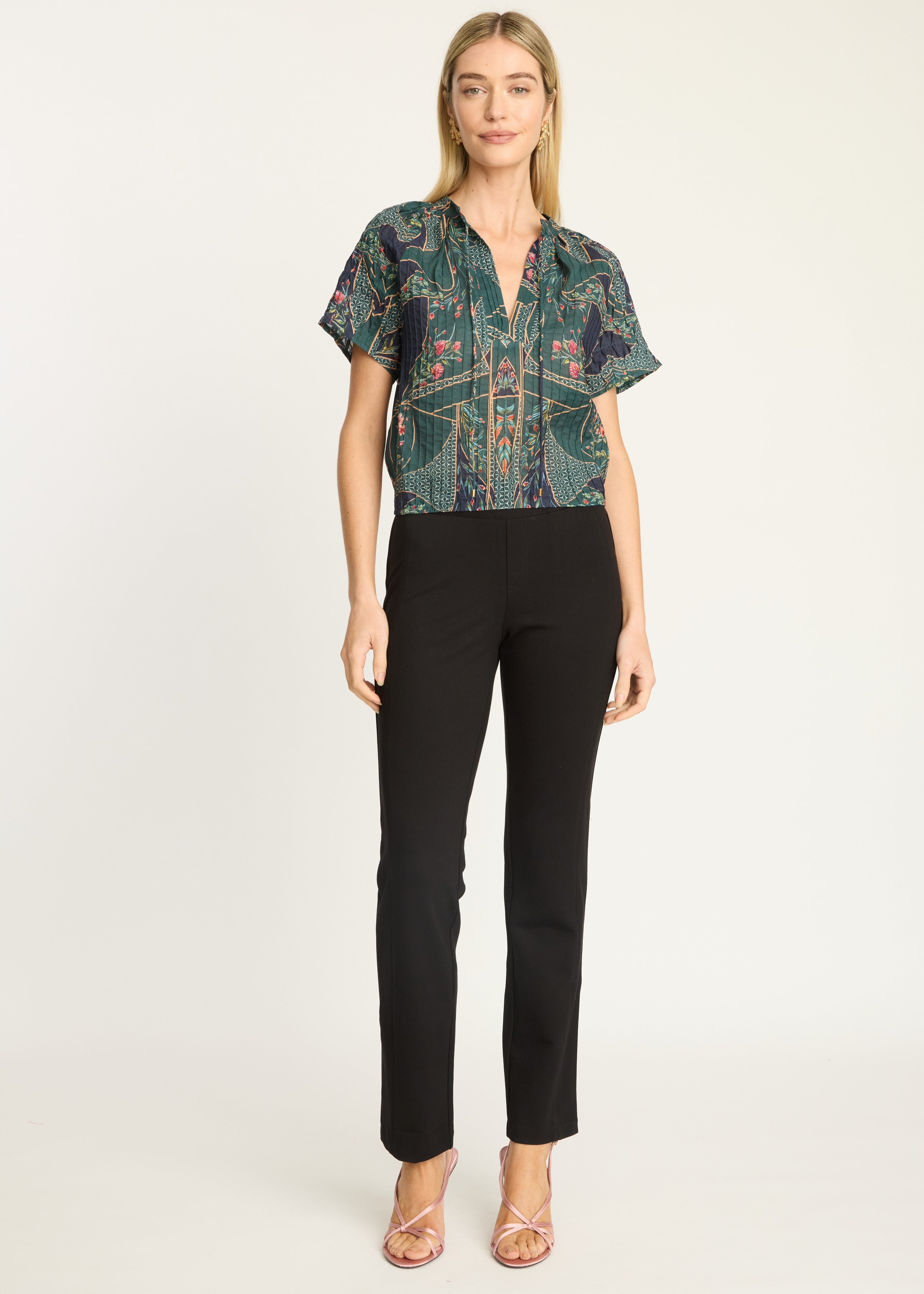 A woman modeling a short-sleeve button-up blouse with a green and black abstract pattern, paired with black cropped pants and metallic sandals, against a plain light gray studio background.