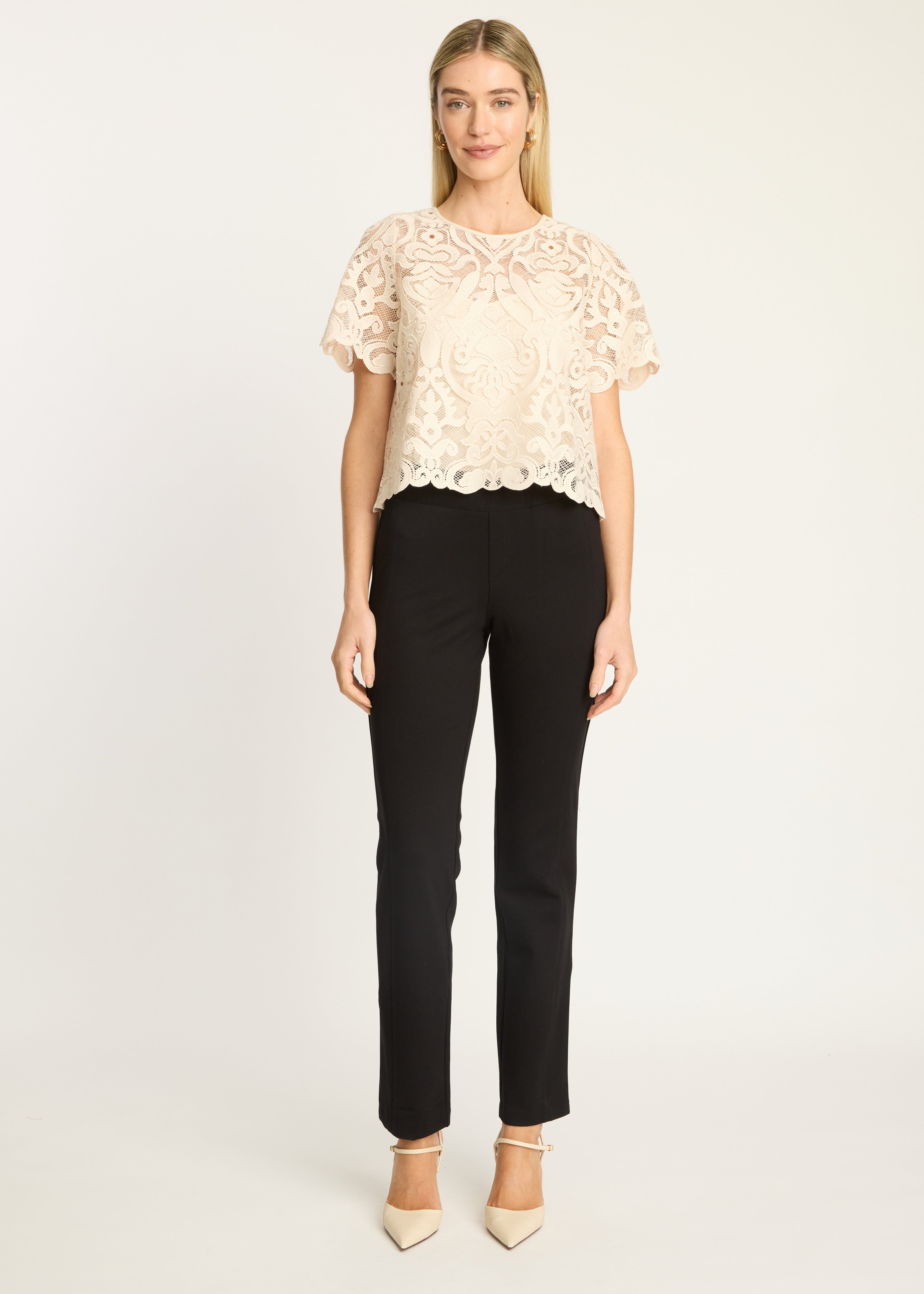 A woman modeling a cream lace short-sleeve top with black trousers and beige heels, standing against a plain light gray studio background.