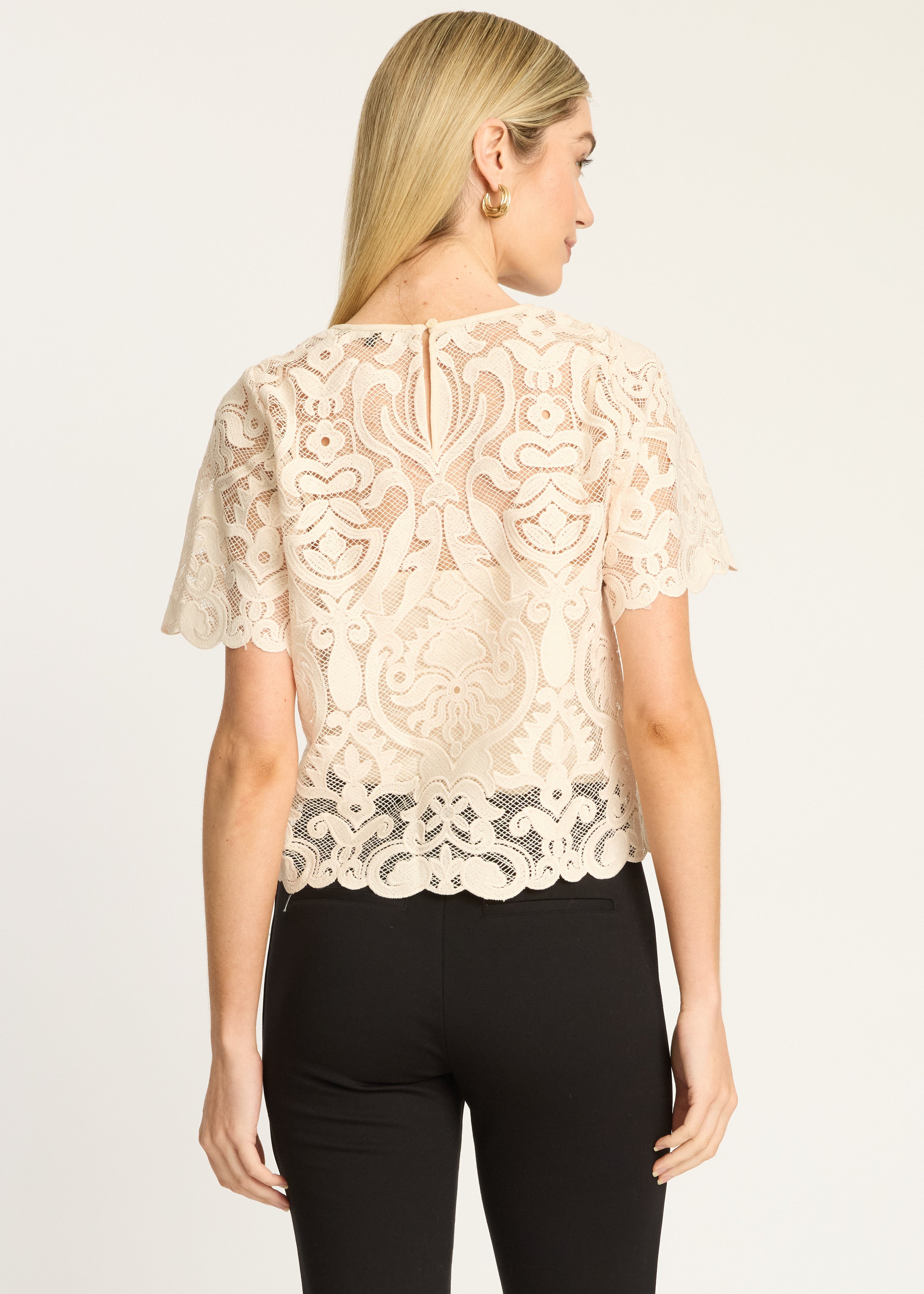 A woman shown from the back wearing a short-sleeve cream lace top with a scalloped hem and black pants, against a plain light background.