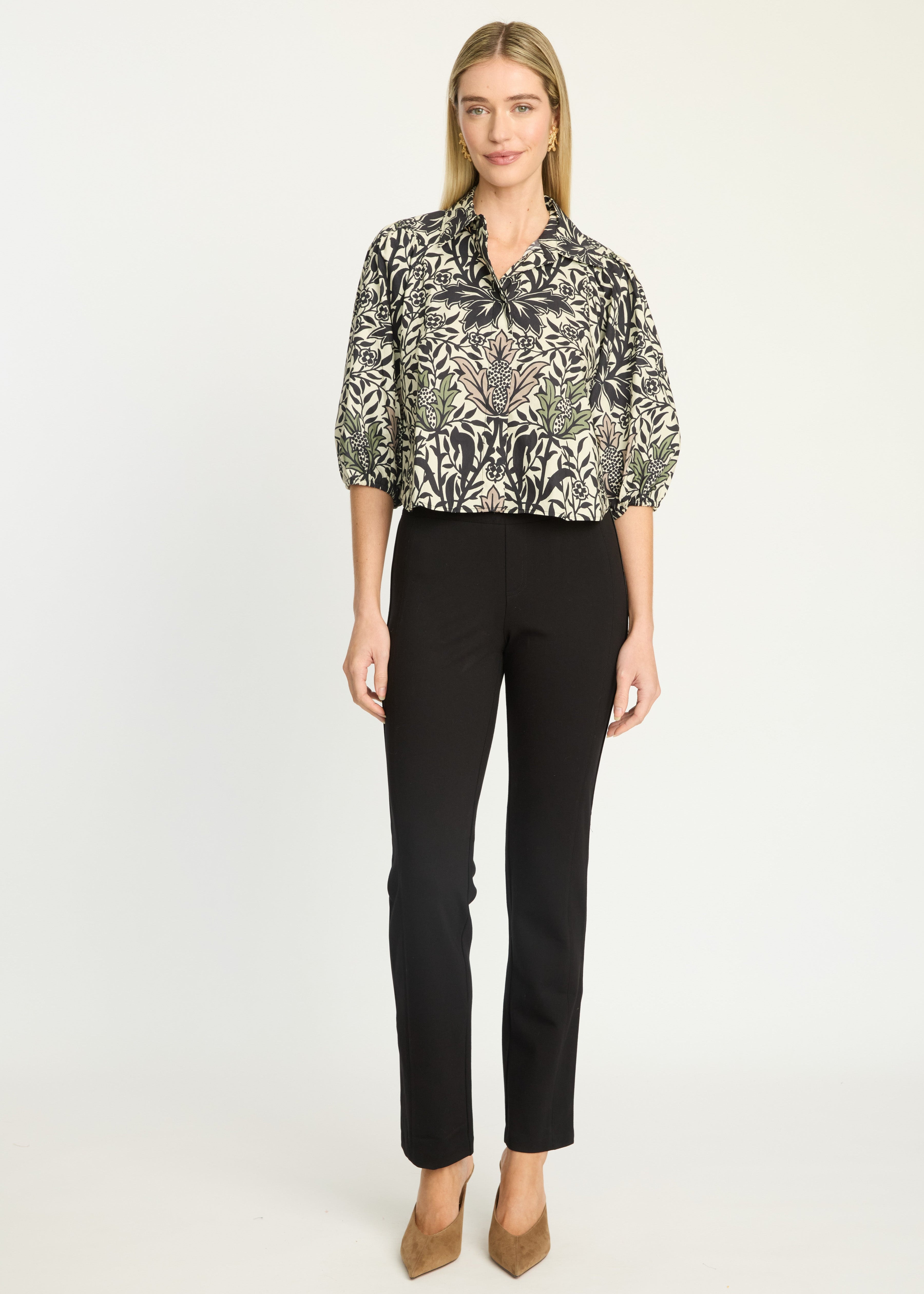 A woman modeling a black and beige botanical print blouse with three-quarter sleeves, paired with black trousers and tan heels, against a plain white studio background.