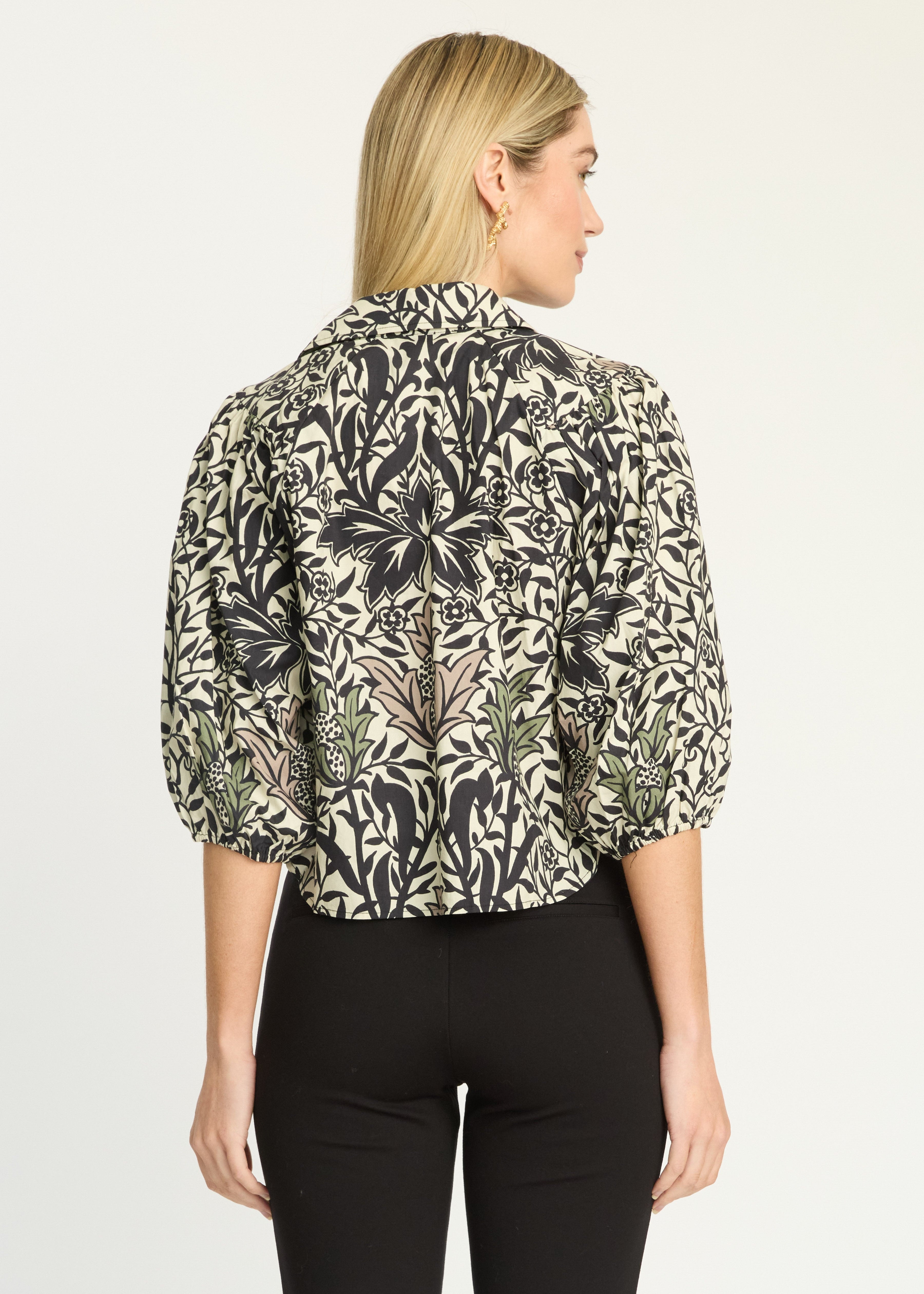 A woman shown from the back wearing a short-sleeve blouse with a black and beige botanical floral pattern, paired with black pants, against a plain light background.