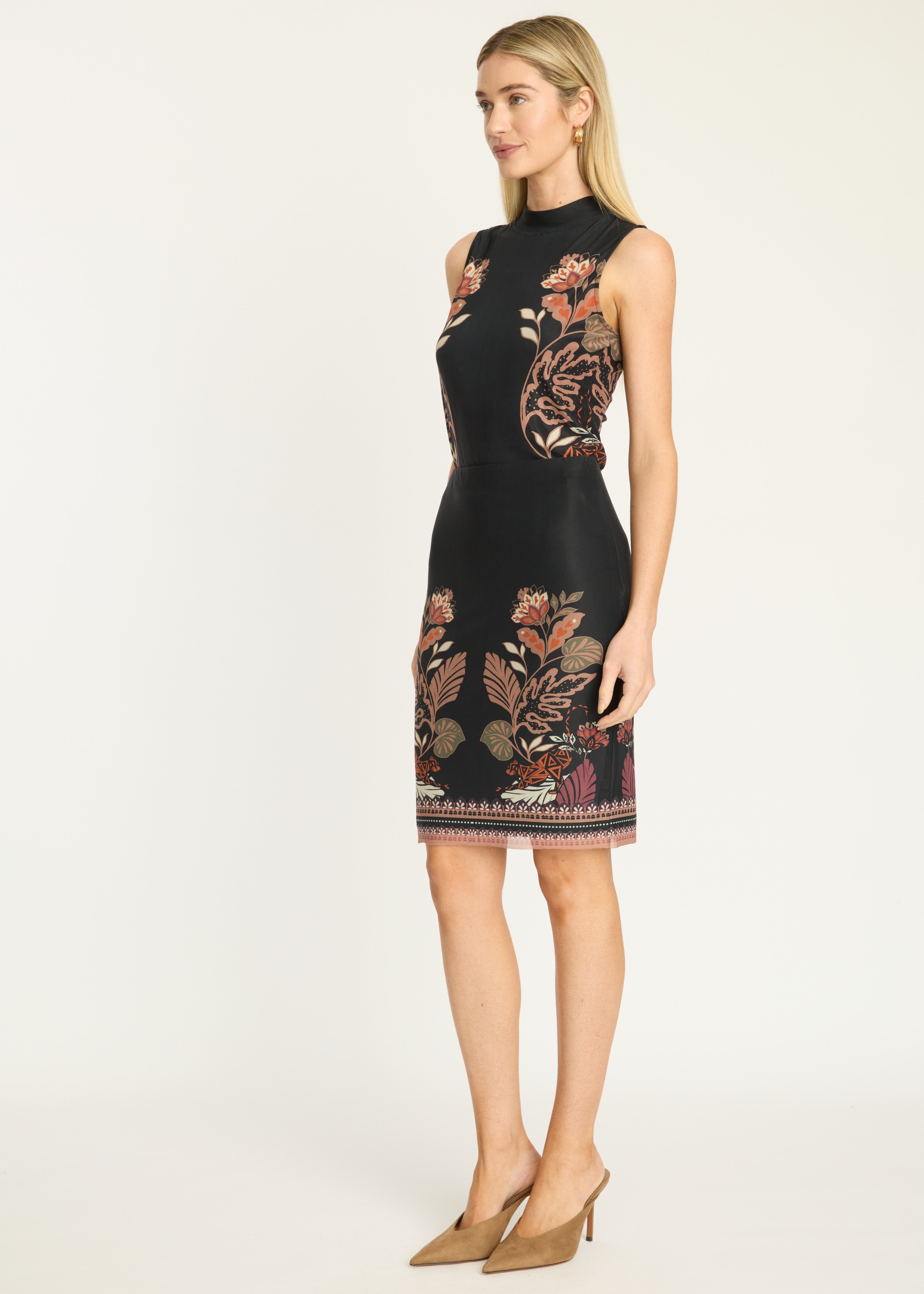A woman modeling a sleeveless black knee-length dress with floral and botanical embroidery in warm tones, paired with tan pointed-toe heels, against a plain light gray studio background.