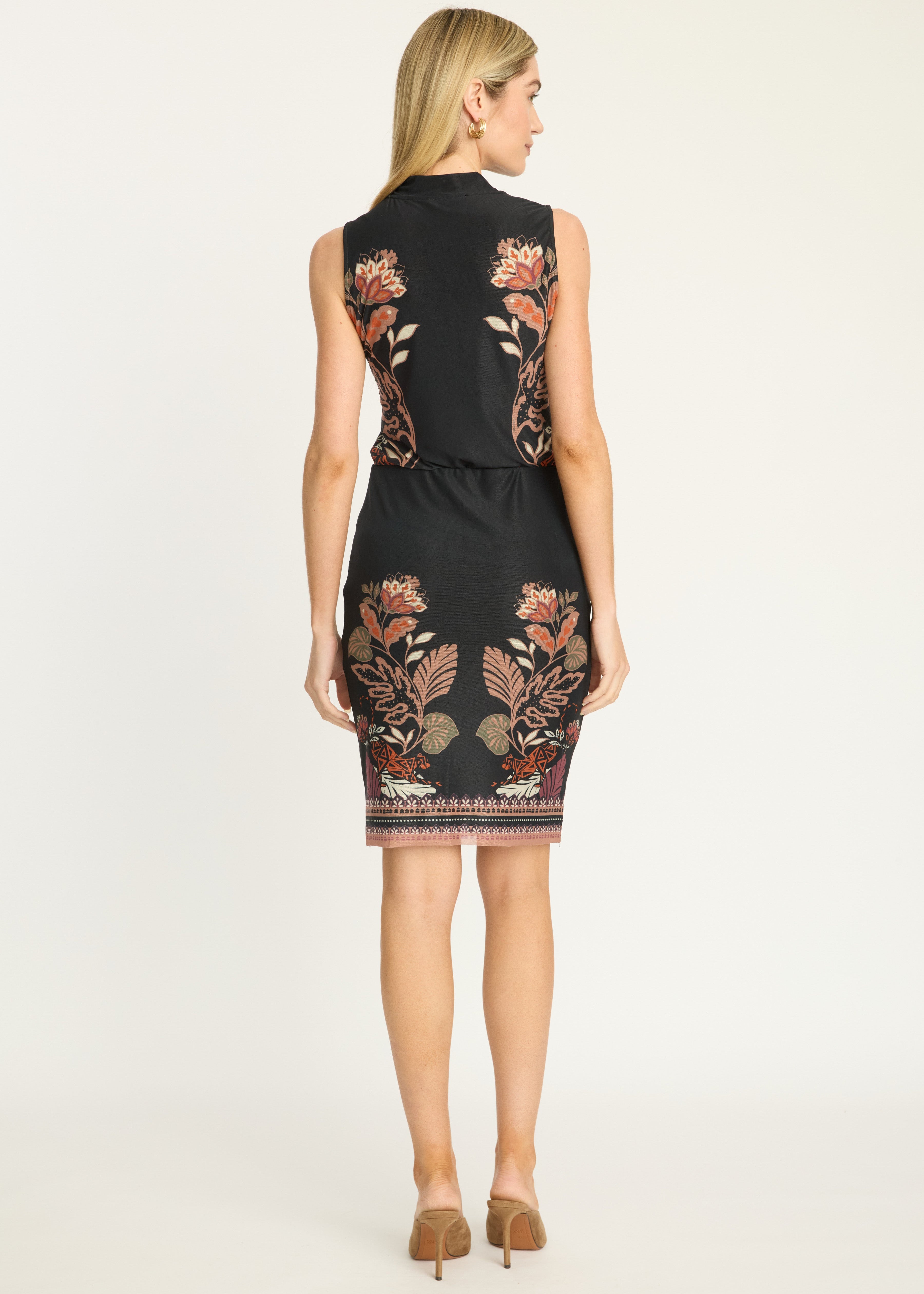 A woman viewed from the back wearing a sleeveless black knee-length dress with floral embroidery in warm tones, paired with tan heels, against a plain light background.