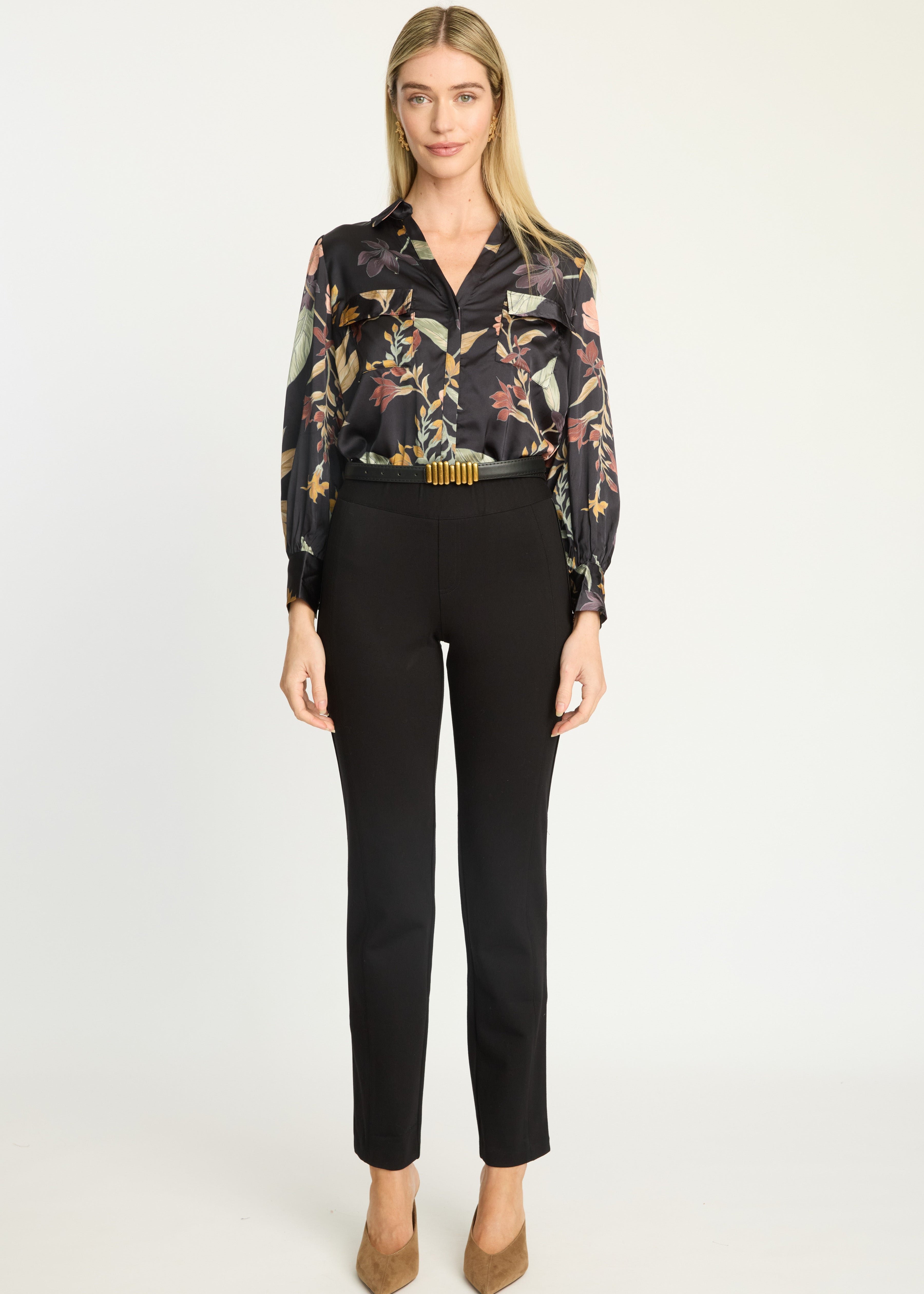 A woman stands against a plain light background wearing a black long-sleeve blouse with a multicolor floral print, black cropped trousers, and tan heels.