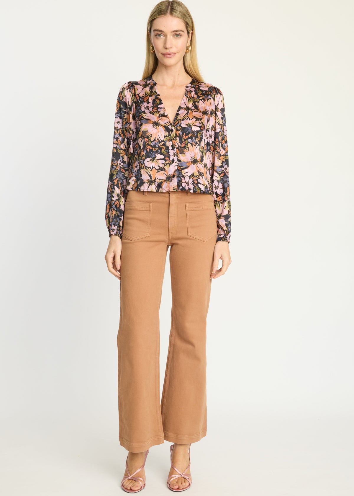 A woman modeling a long-sleeve floral button-up blouse with a V-neck and wide cuffs, paired with tan cropped flare pants and strappy heels, against a plain light gray studio background.