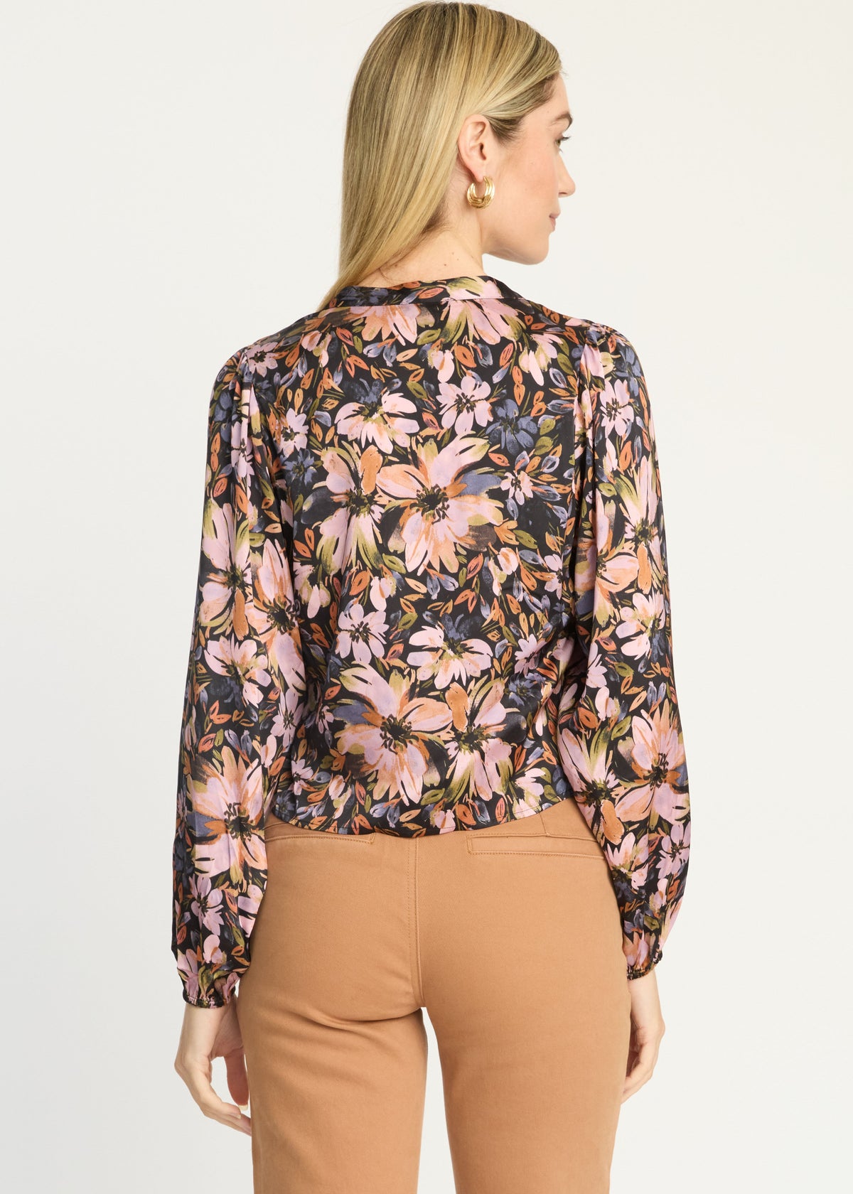 A woman shown from the back wearing a long-sleeve floral blouse with a dark base and pink, peach, and green floral print, paired with tan pants, against a plain light background.