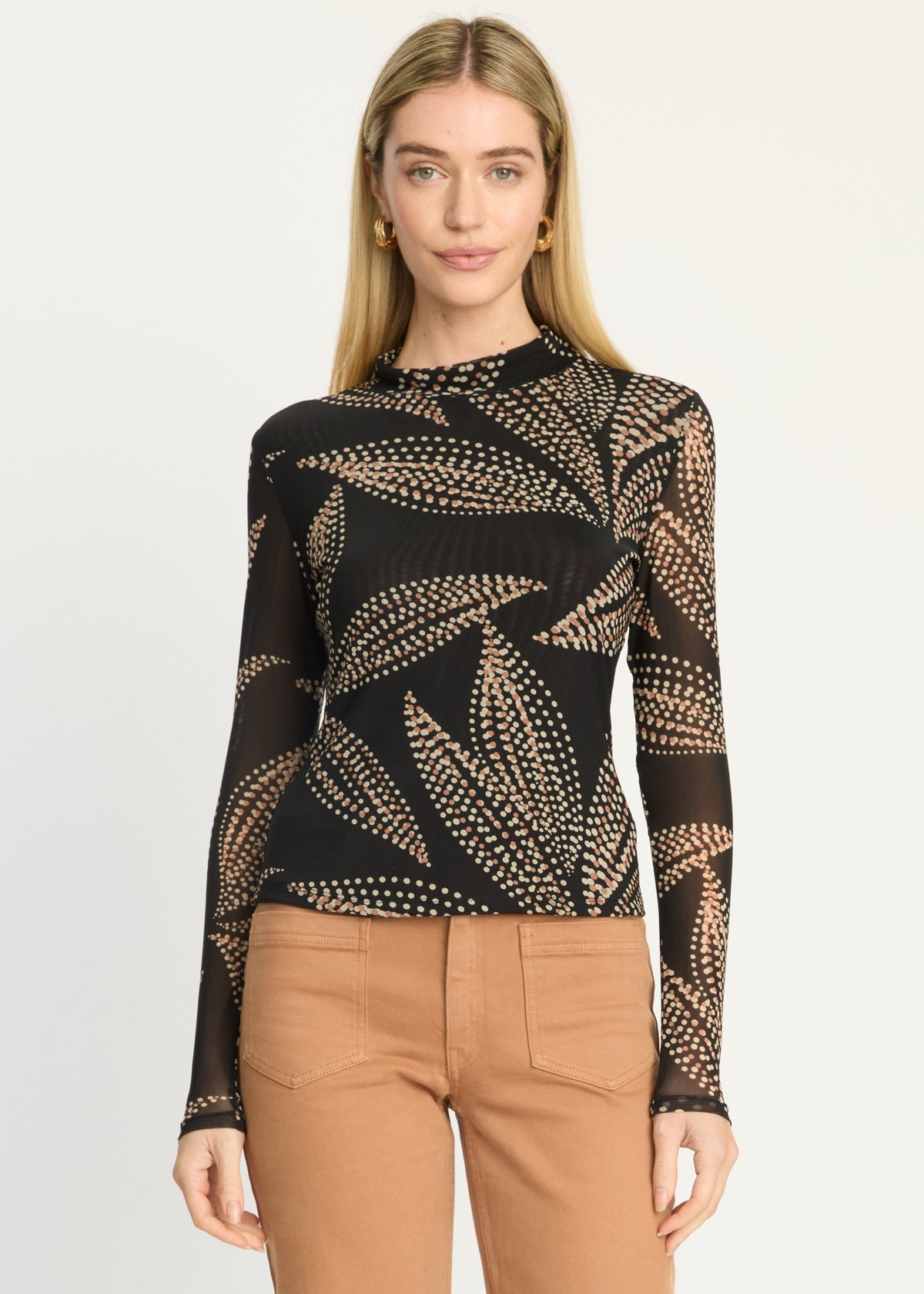A woman modeling a black long-sleeve knit top with a beige leaf pattern, paired with tan pants, against a plain light gray background.