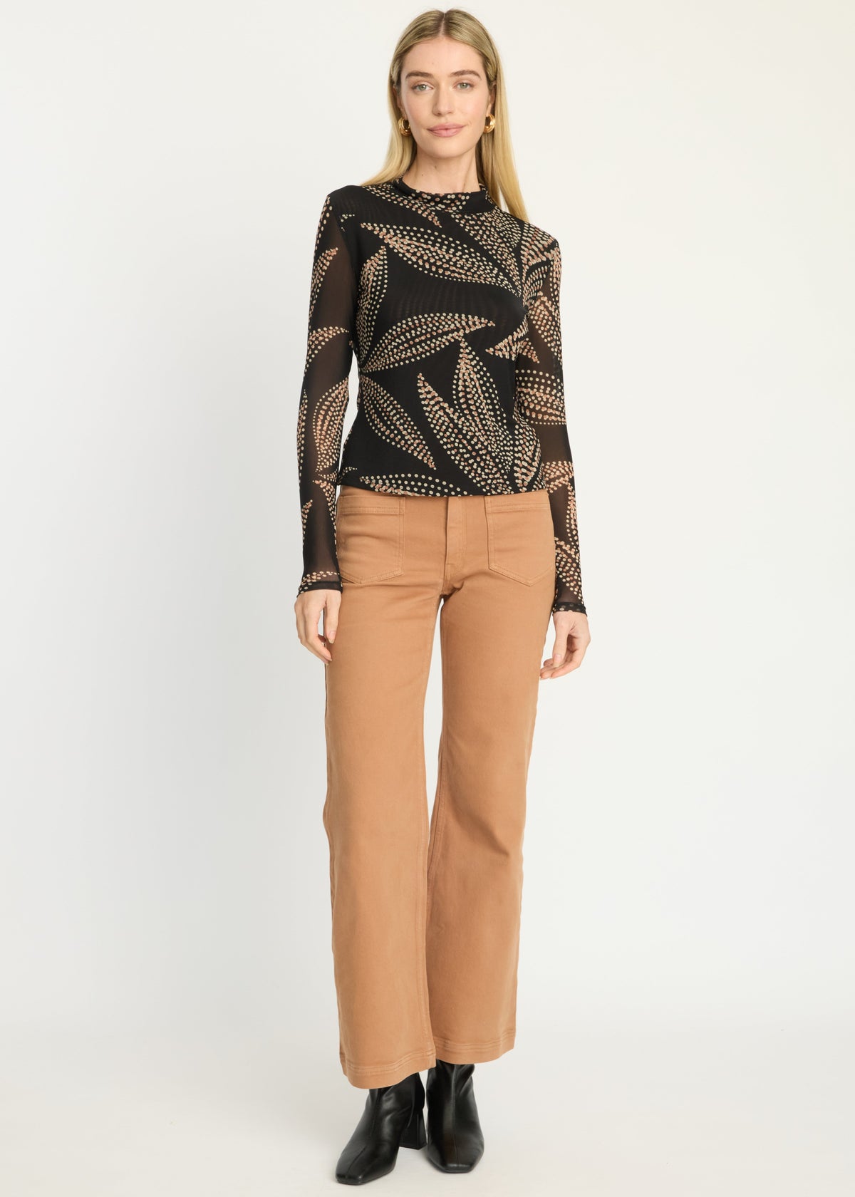 A woman stands against a plain light gray studio background wearing a black long-sleeve top with a beige leaf pattern, tan straight-leg pants, and black ankle boots.