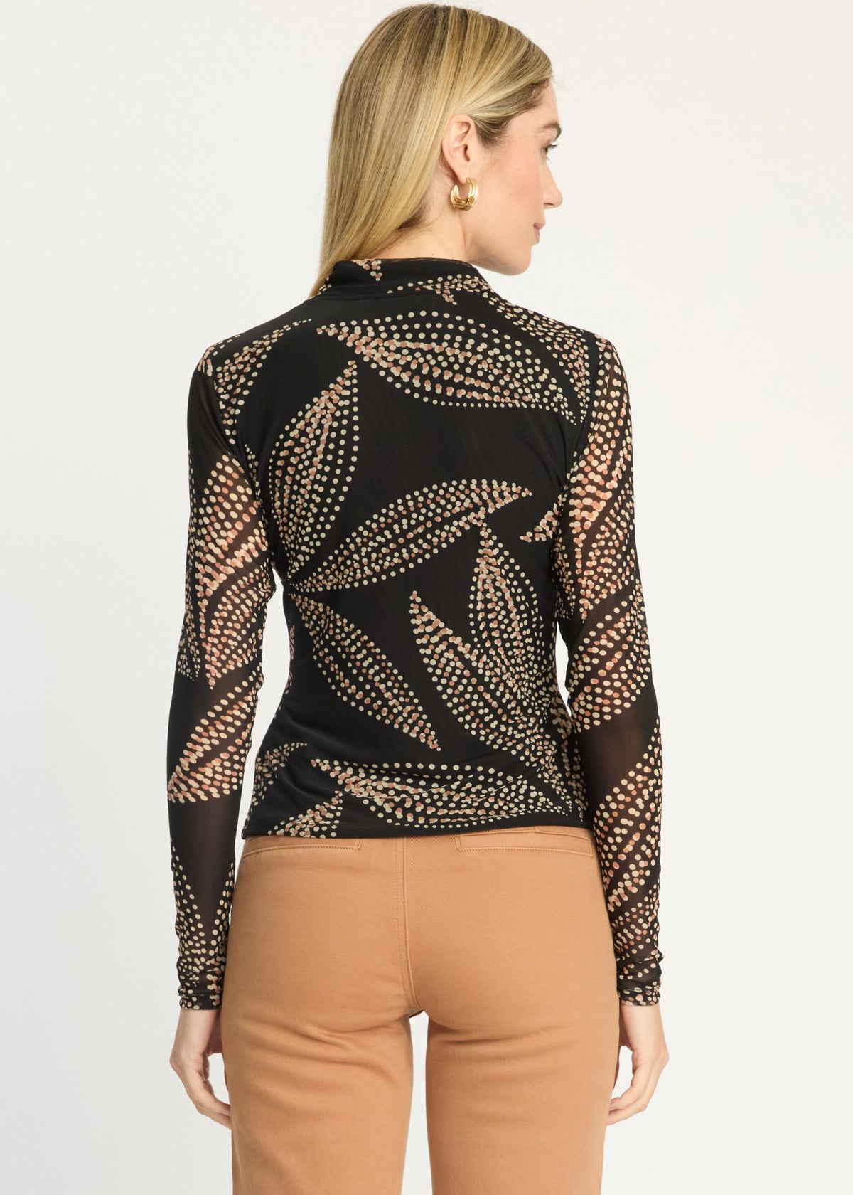 A woman shown from the back wearing a black long-sleeve top with a sheer mesh leaf pattern outlined in gold beads, paired with tan pants, against a plain white studio background.