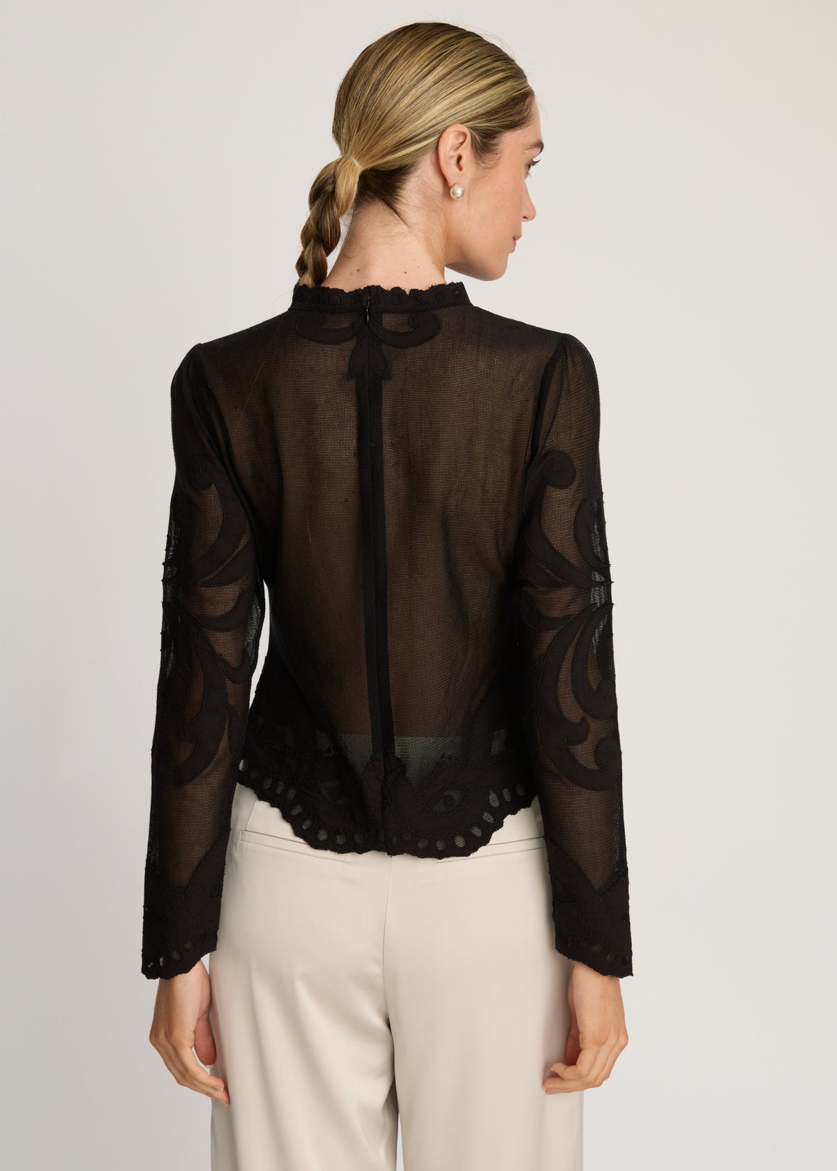 Woman wearing a black sheer blouse with long sleeves and white pants on a light gray background