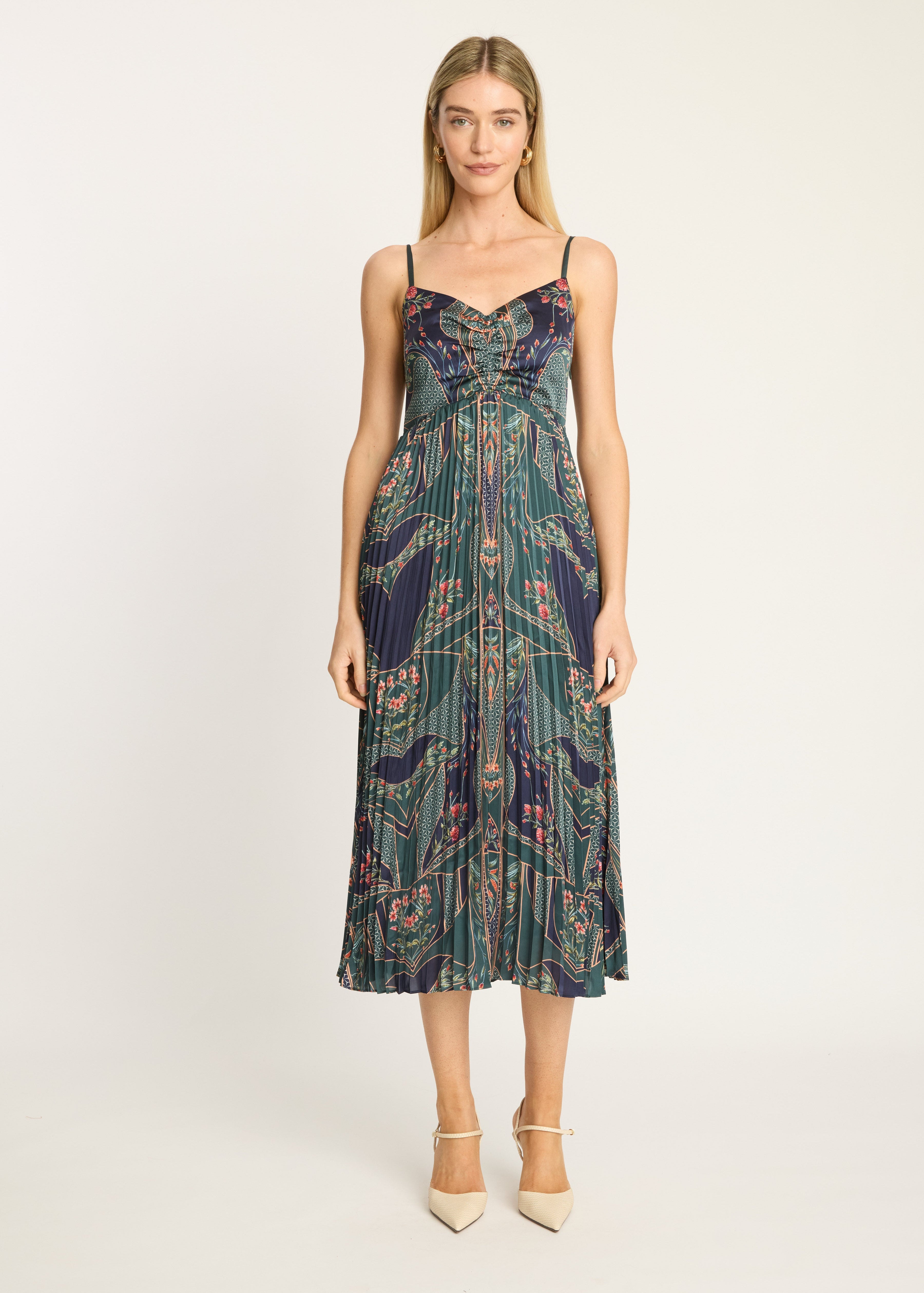 A woman modeling a sleeveless midi dress with thin straps, a fitted bodice, and a pleated skirt. The dress features an intricate abstract pattern in dark teal, navy, and muted pink.
