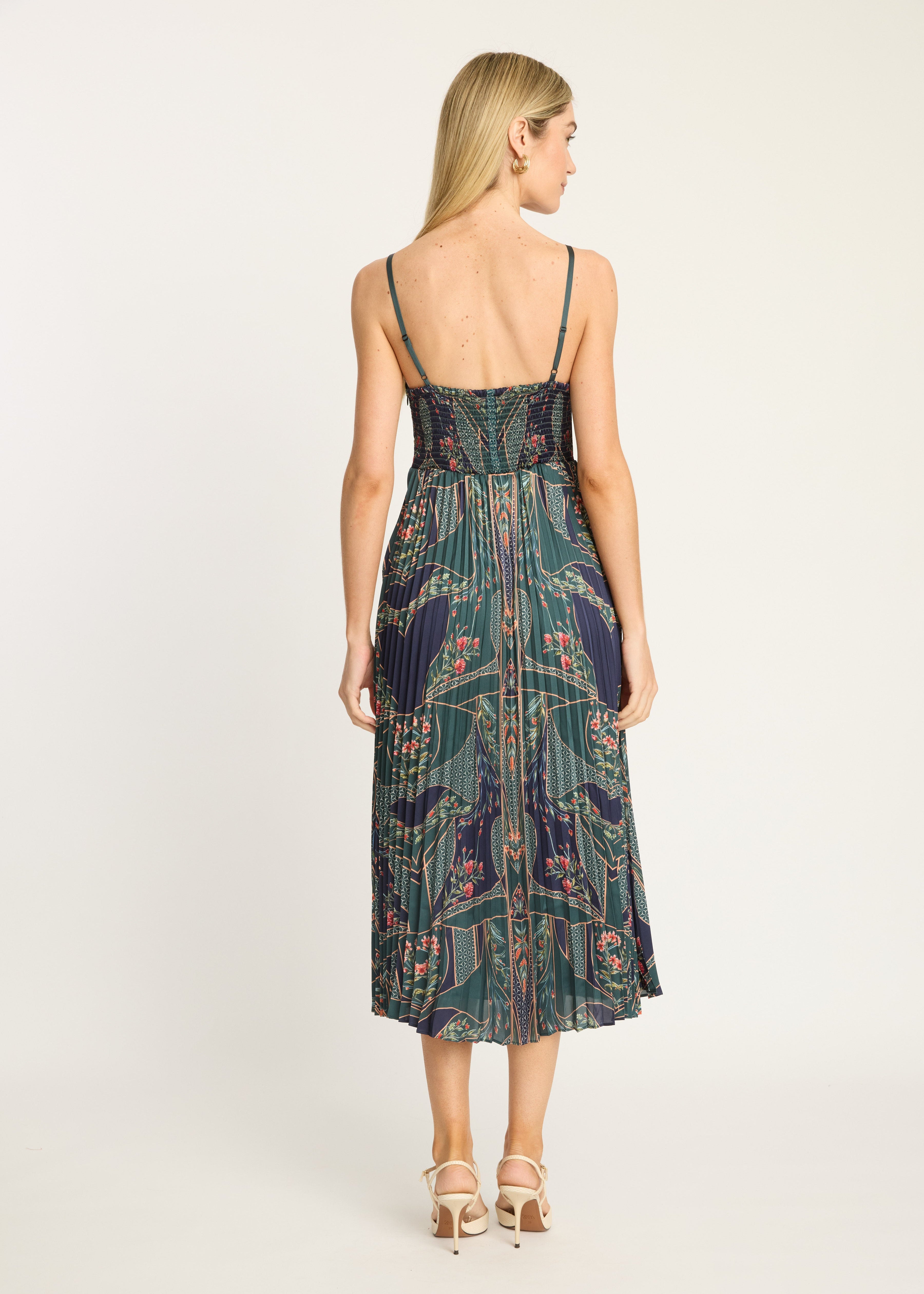 A woman viewed from the back wearing a sleeveless midi dress with thin straps, a fitted bodice, and a flowing printed skirt. The dress has an abstract botanical pattern in dark teal, green, navy, and burgundy tones.