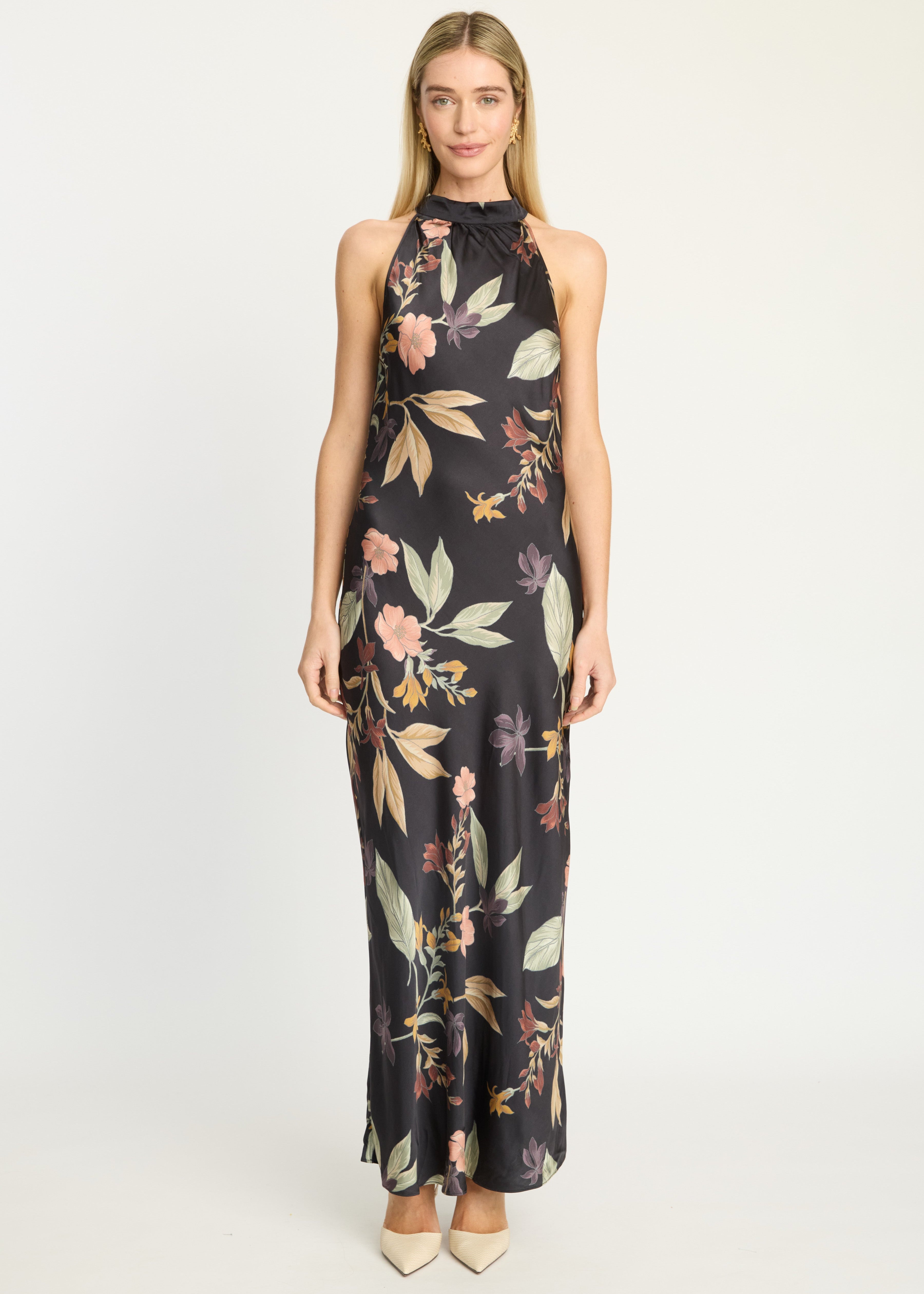 A woman modeling a sleeveless, high-neck maxi dress with a black satin-like base and a multicolor floral and leaf print, standing against a plain light gray studio background.