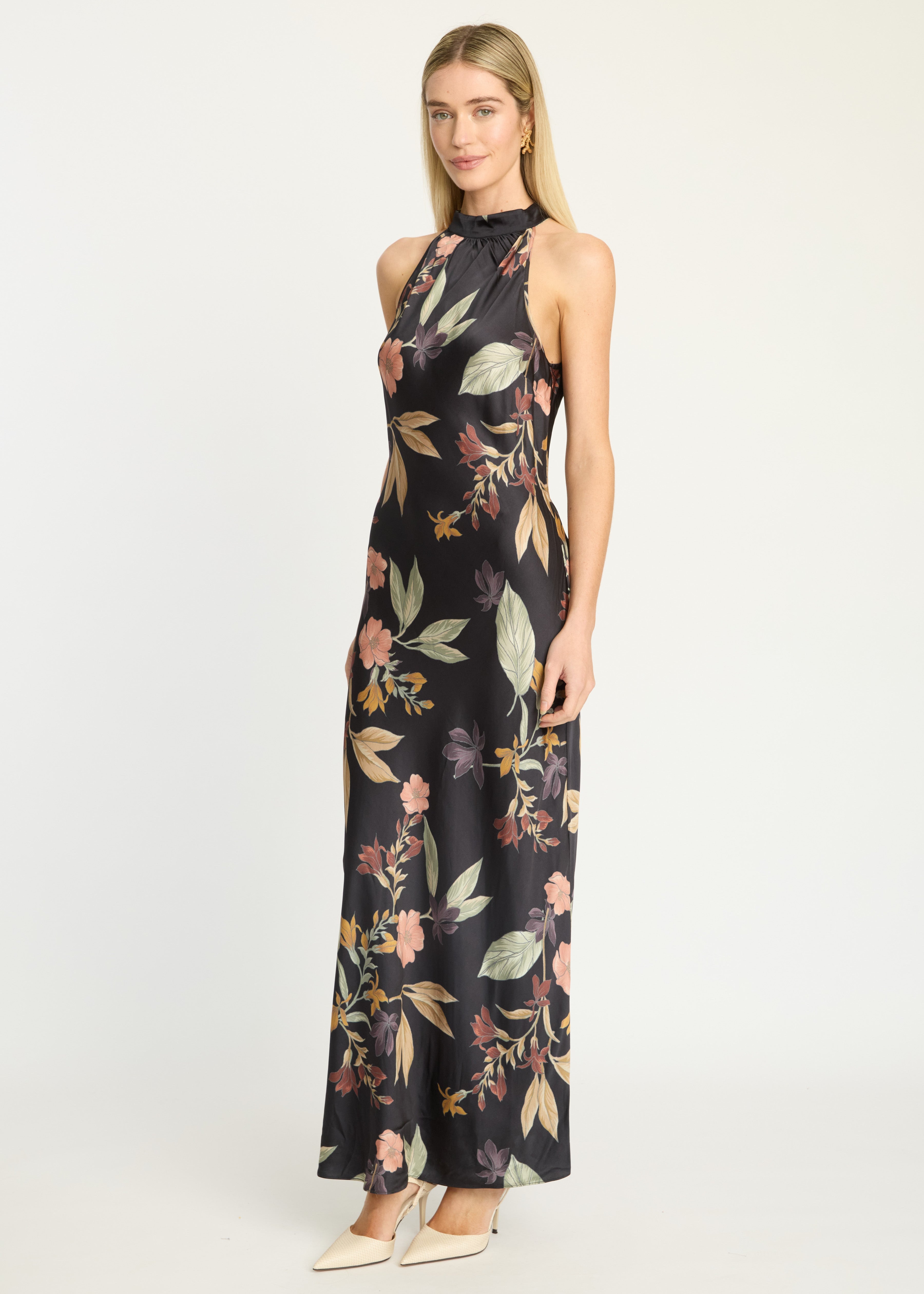 A woman modeling a sleeveless, high-neck, floor-length maxi dress with a black base and a multicolor floral print in pink, green, and beige, paired with white pointed-toe heels against a plain white studio background.