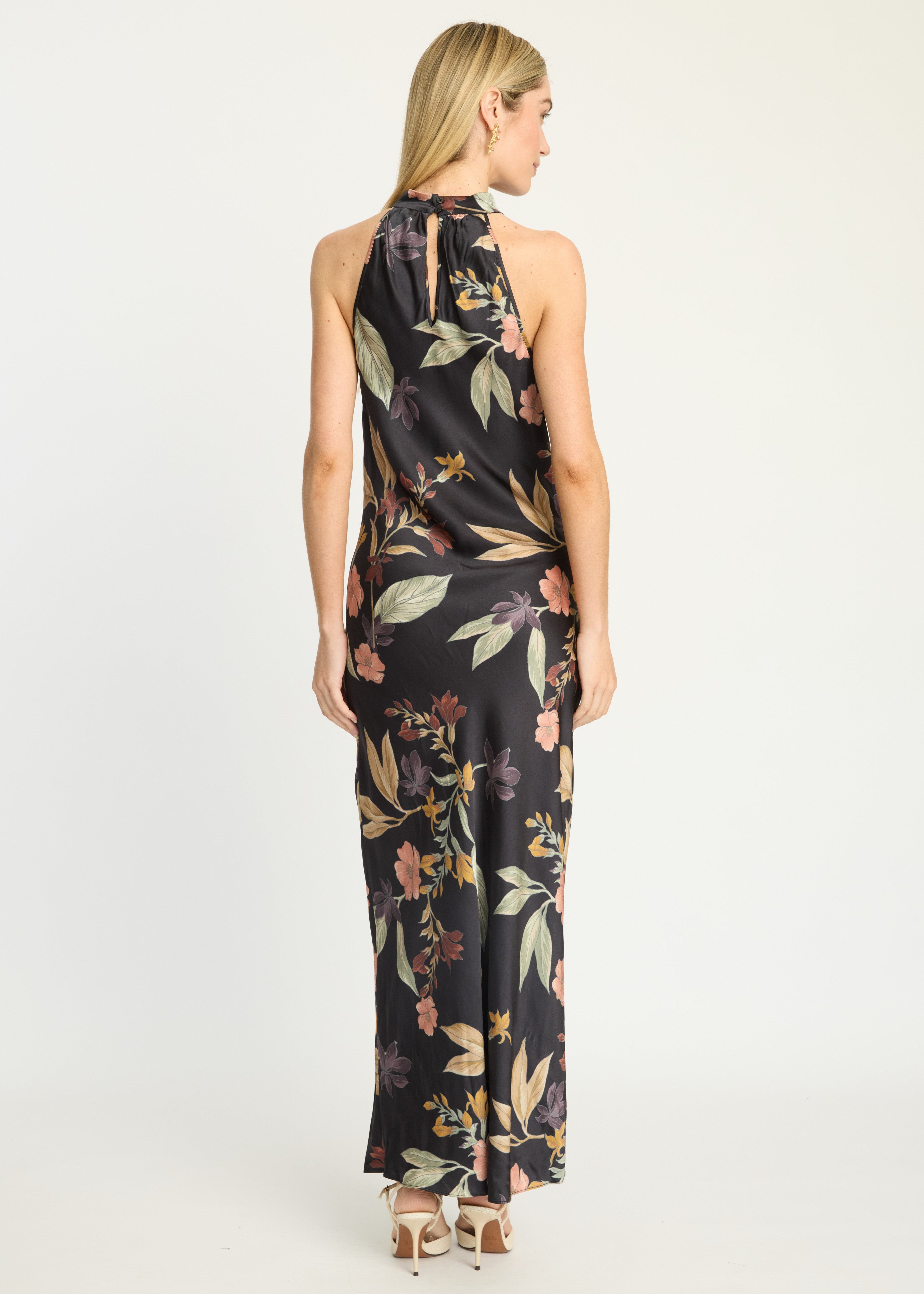 A woman viewed from the back wearing a sleeveless halter-neck maxi dress with a black base and a multicolor floral print featuring green, pink, and orange tones, paired with beige high-heeled sandals against a plain white studio background.