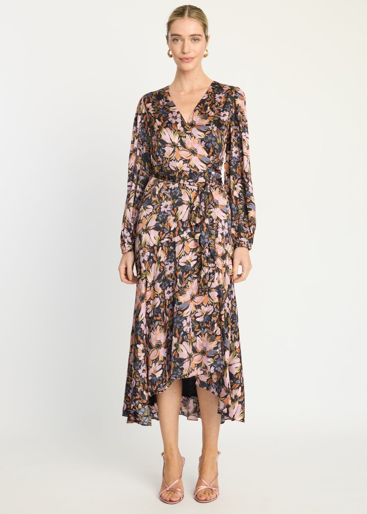 A woman modeling a long-sleeve floral midi dress with a wrap-style V-neck and high-low hem, paired with strappy heels, against a plain light gray studio background.