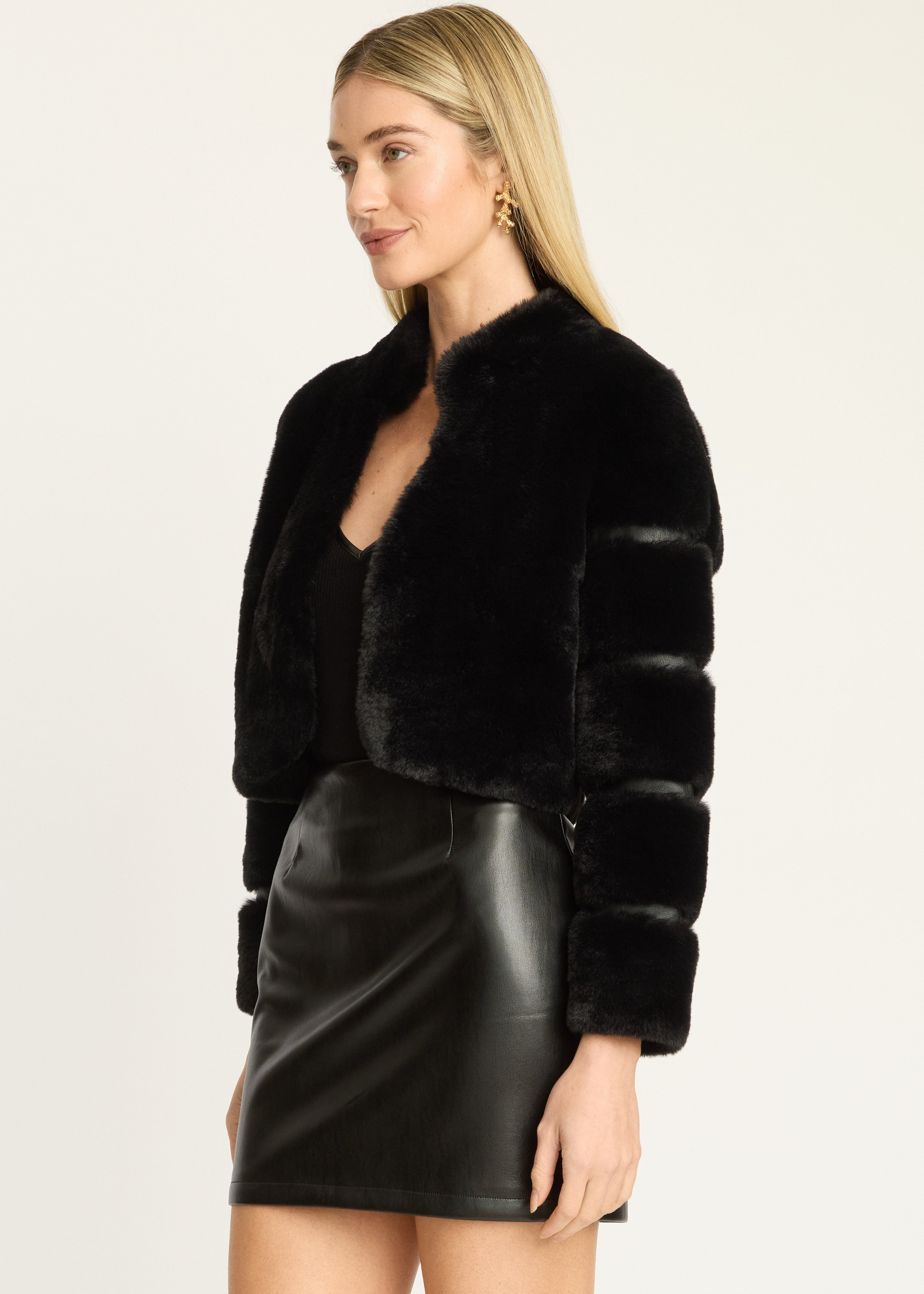 A blonde model wearing a black cropped faux fur jacket with puffed sleeves and a black faux leather mini skirt, photographed against a plain light background.