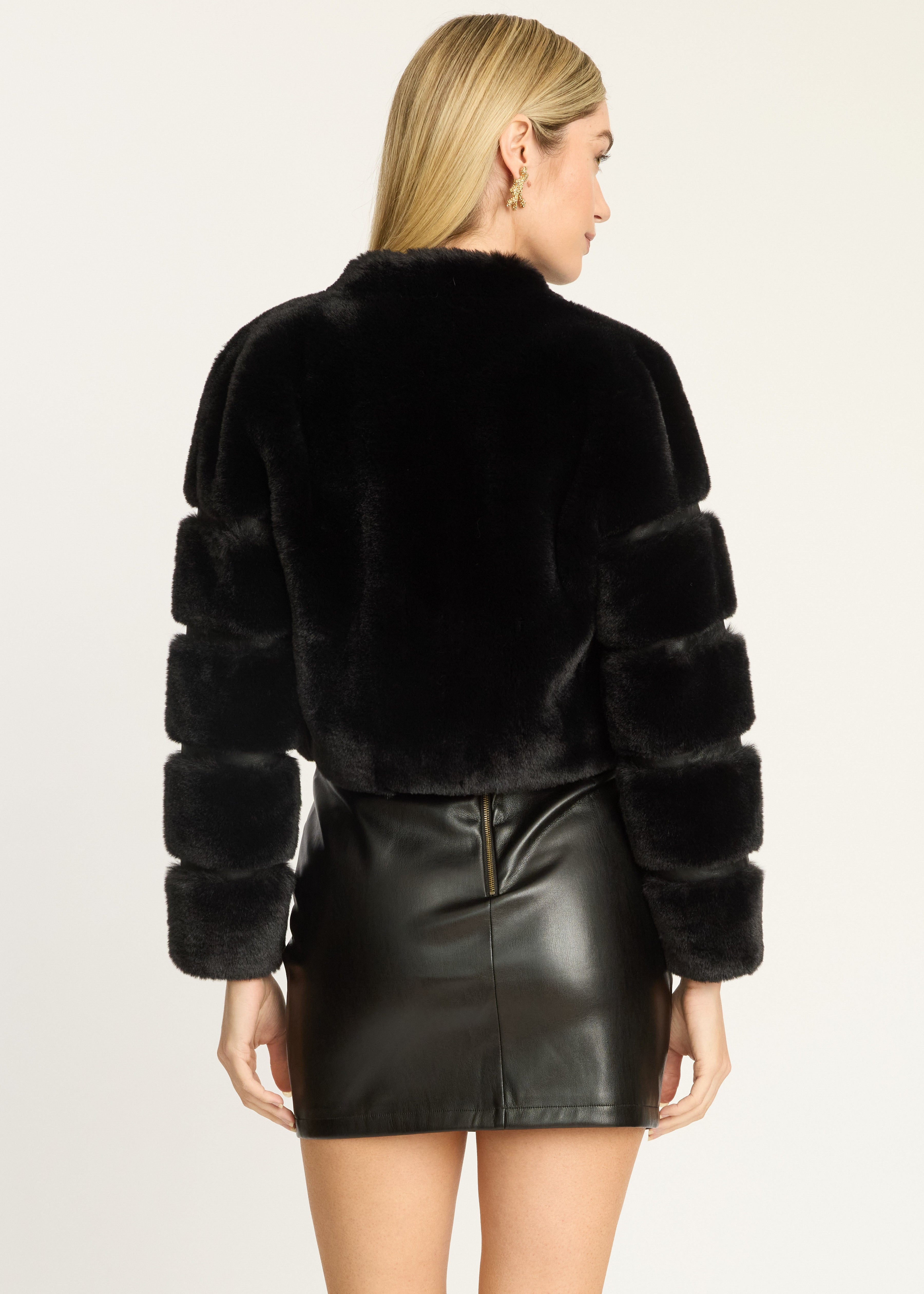 A woman shown from the back wearing a black cropped faux-fur jacket with ribbed sleeves and a black faux-leather mini skirt, against a plain light background.