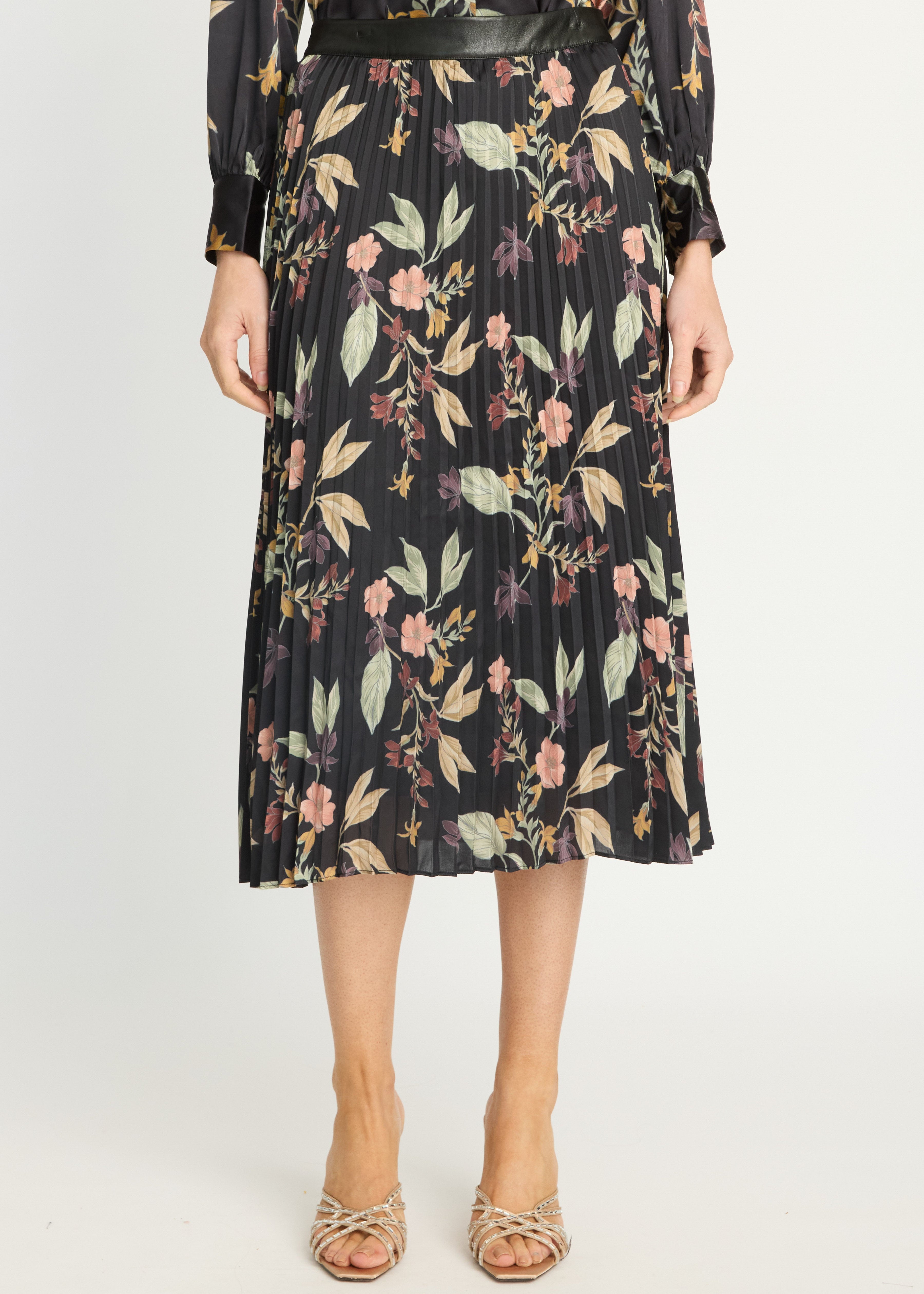 A model wearing a black pleated midi skirt with a floral print in pink, green, and beige, styled with a black belt and beige strappy heels against a white studio background.