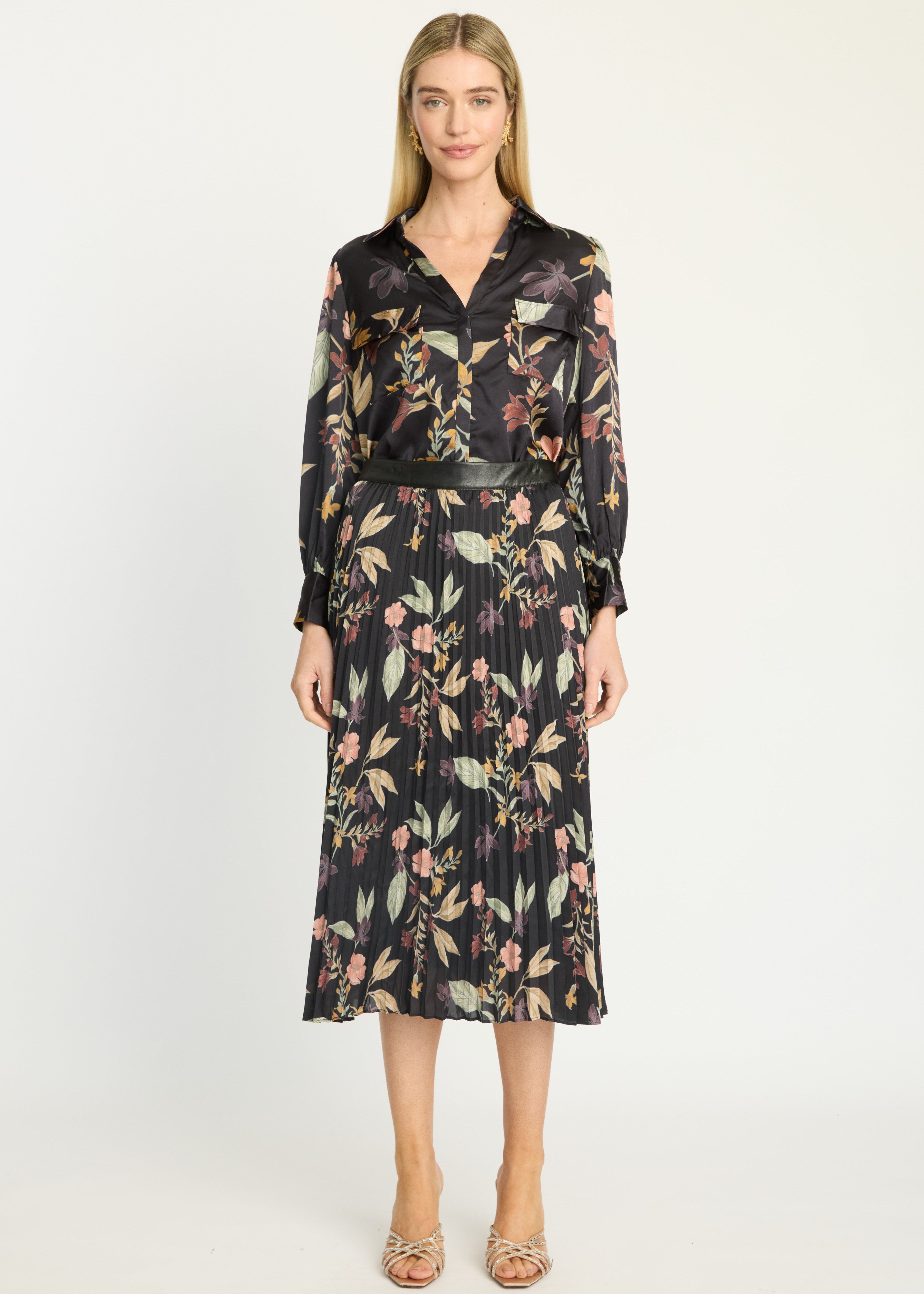 A woman modeling a black long-sleeve midi shirt dress with a floral print in pink, green, and beige, paired with metallic heeled sandals, against a plain white studio background.