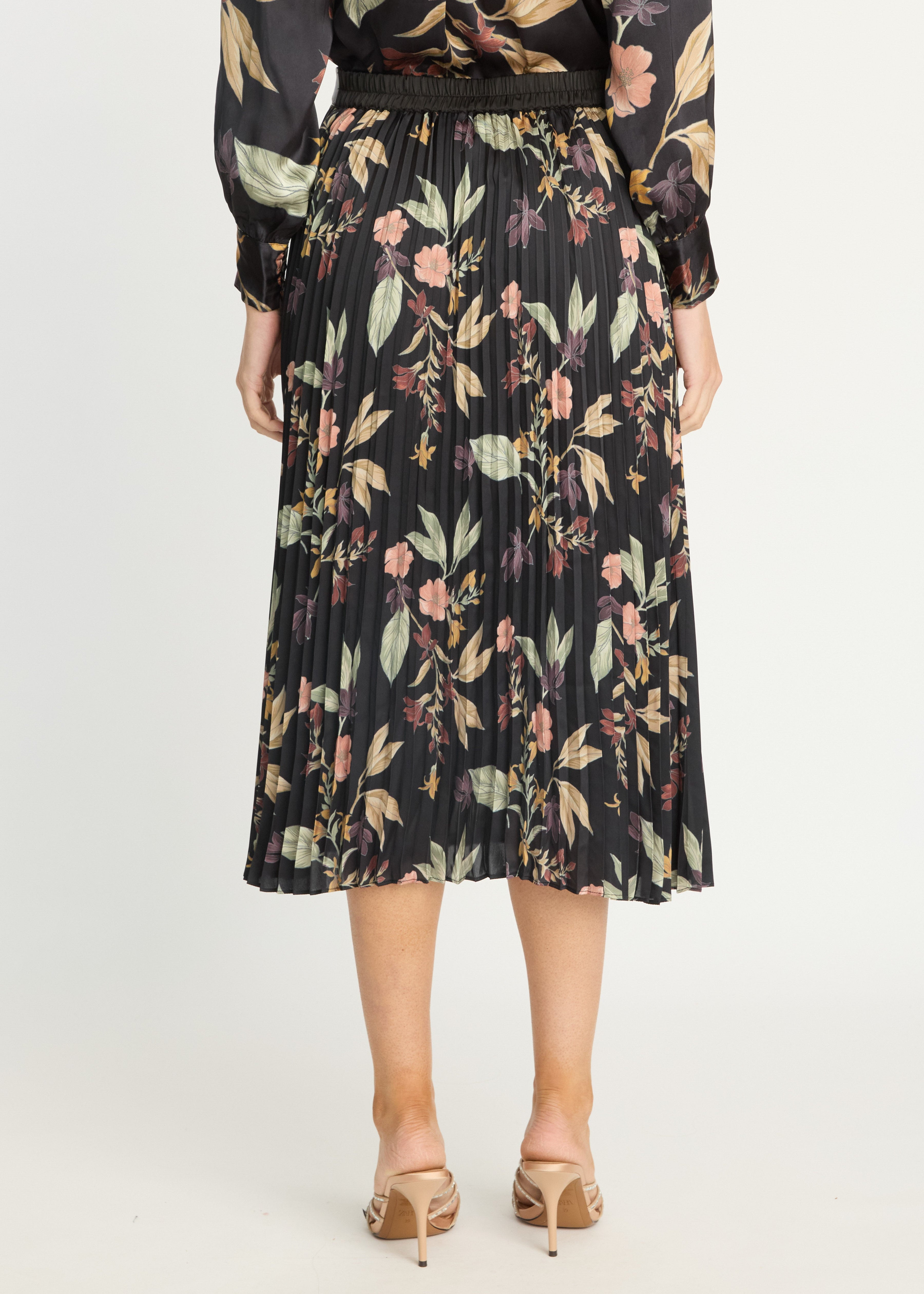 A woman is shown from the back wearing a black pleated midi skirt with a multicolor floral print, paired with a matching long-sleeve top and beige heeled sandals, against a plain light gray studio background.