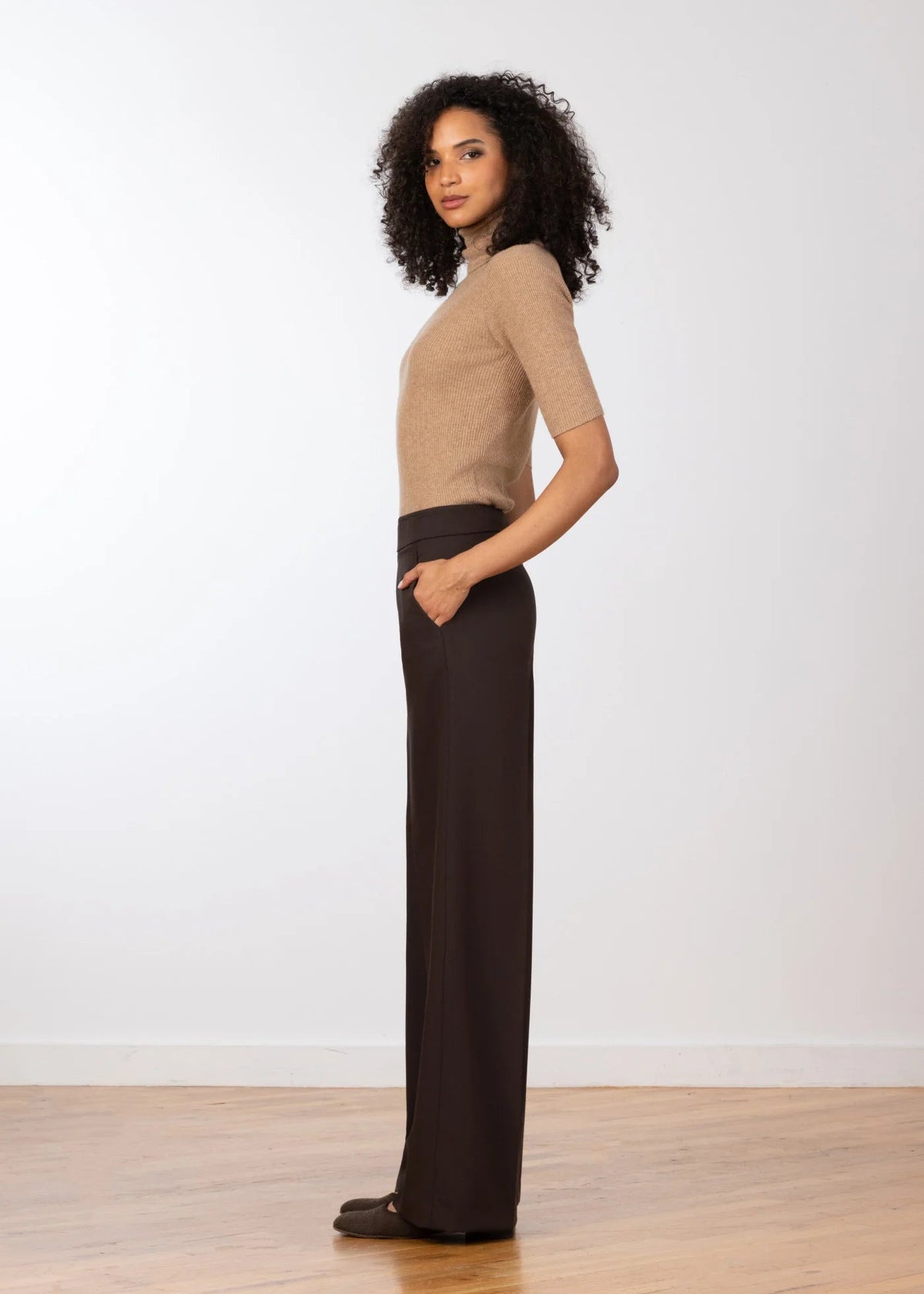 A woman with curly black hair stands in profile wearing a beige short-sleeve mock-neck top and dark brown wide-leg trousers, standing on a light wood floor against a white wall.