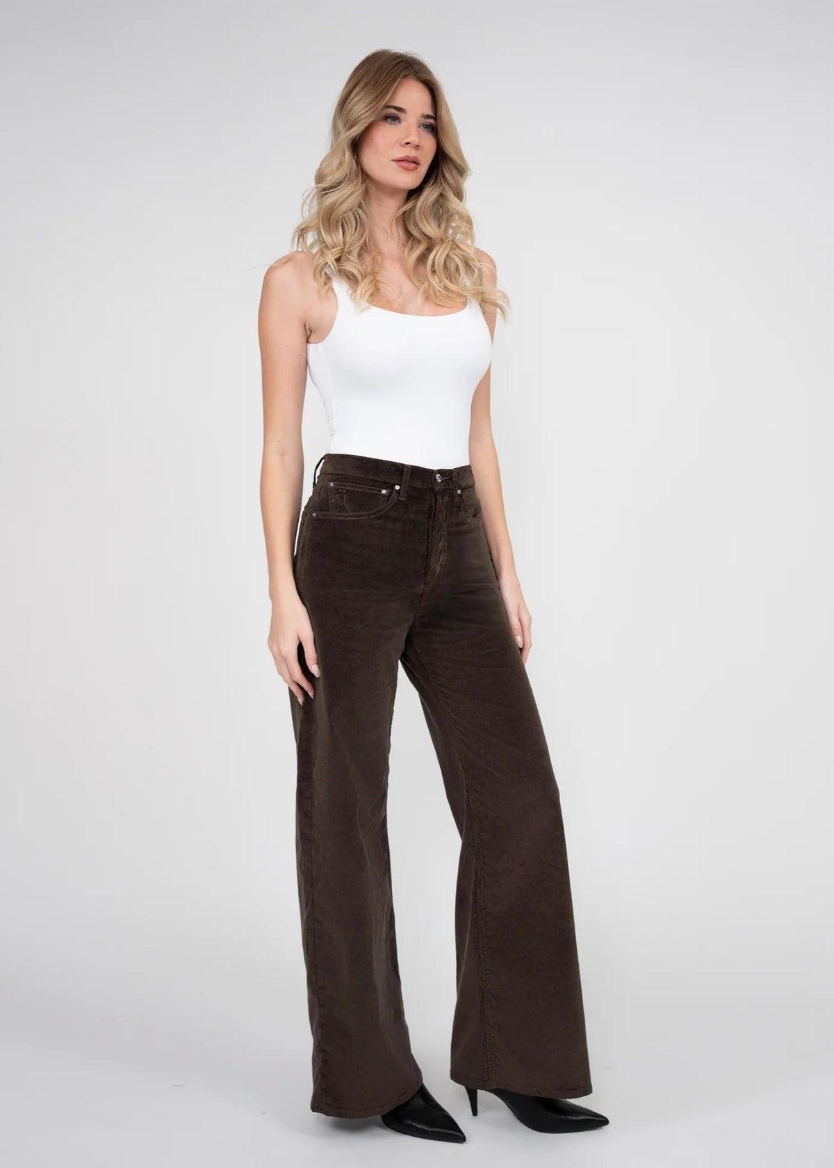 A woman modeling a pair of dark brown wide-leg corduroy pants with a white tank top and black heels, standing against a light gray studio background.