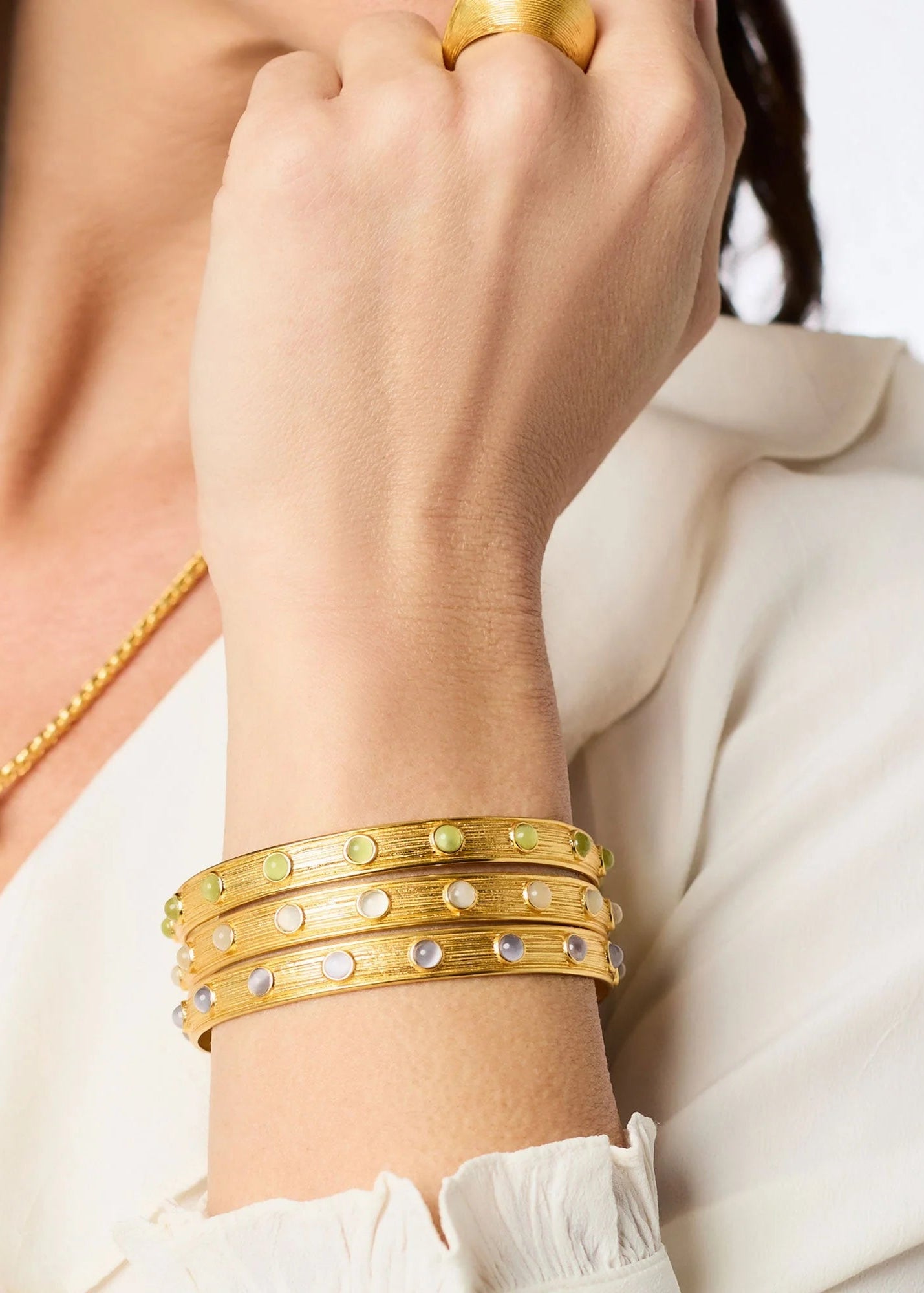 A close-up of a person wearing a stack of three gold bangle bracelets on their wrist, each decorated with small round green and white stones. The person is also wearing a gold necklace and a gold ring, and is dressed in a white blouse.