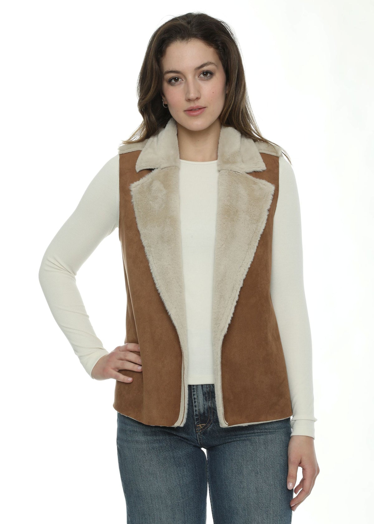 A woman modeling a brown suede shearling vest with a cream-colored collar and lining, worn over a cream long-sleeve top and blue jeans against a white studio background.