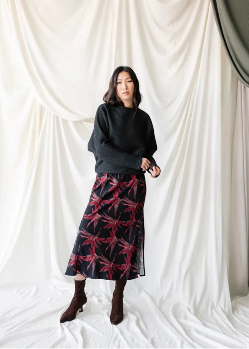 A woman wearing a dark charcoal gray knit sweater and a black skirt with a red and pink tropical leaf pattern, standing against a draped white fabric backdrop.