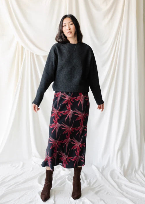 A woman modeling a dark charcoal gray knit sweater paired with a black skirt featuring a red and pink floral pattern, set against a draped white fabric backdrop.