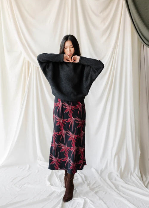 A woman wearing a dark charcoal gray knit sweater with wide batwing sleeves and a black skirt with a red floral pattern, posing in front of a draped white fabric backdrop.