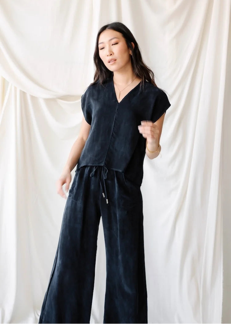 A woman modeling a dark navy blue velvet jumpsuit with a V-neck, short sleeves, and wide-leg pants, standing against a draped white fabric backdrop.