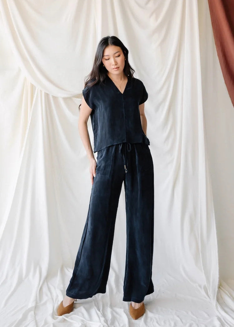 A woman modeling a dark navy two-piece lounge set consisting of a short-sleeve V-neck top and matching wide-leg pants, posed against a draped white fabric backdrop.