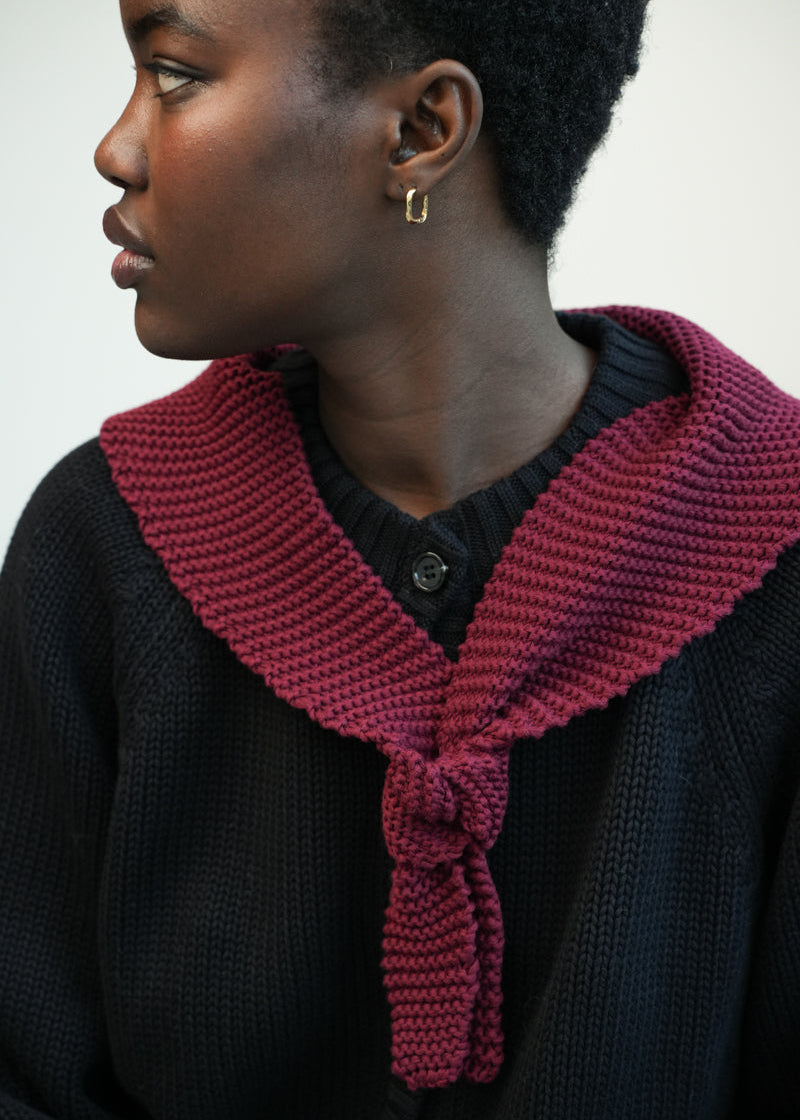 A close-up side profile of a person wearing a dark knit top with a prominent burgundy knitted collar tied at the front, against a plain light background.