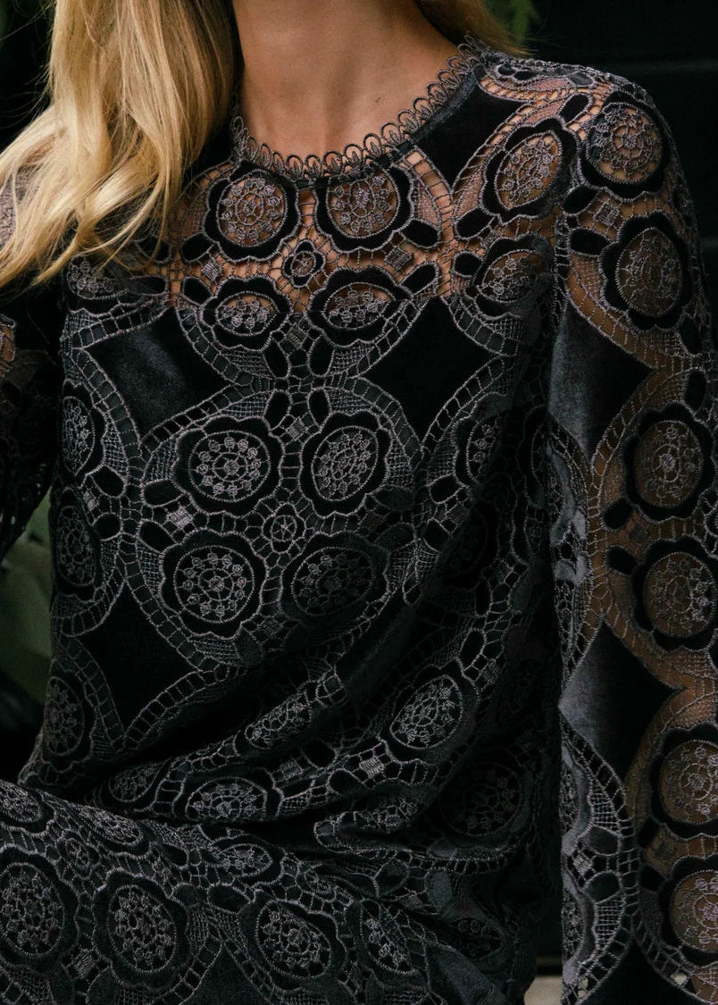 A close-up of a woman wearing a black long-sleeve top with an intricate floral lace overlay and sheer mesh pattern.